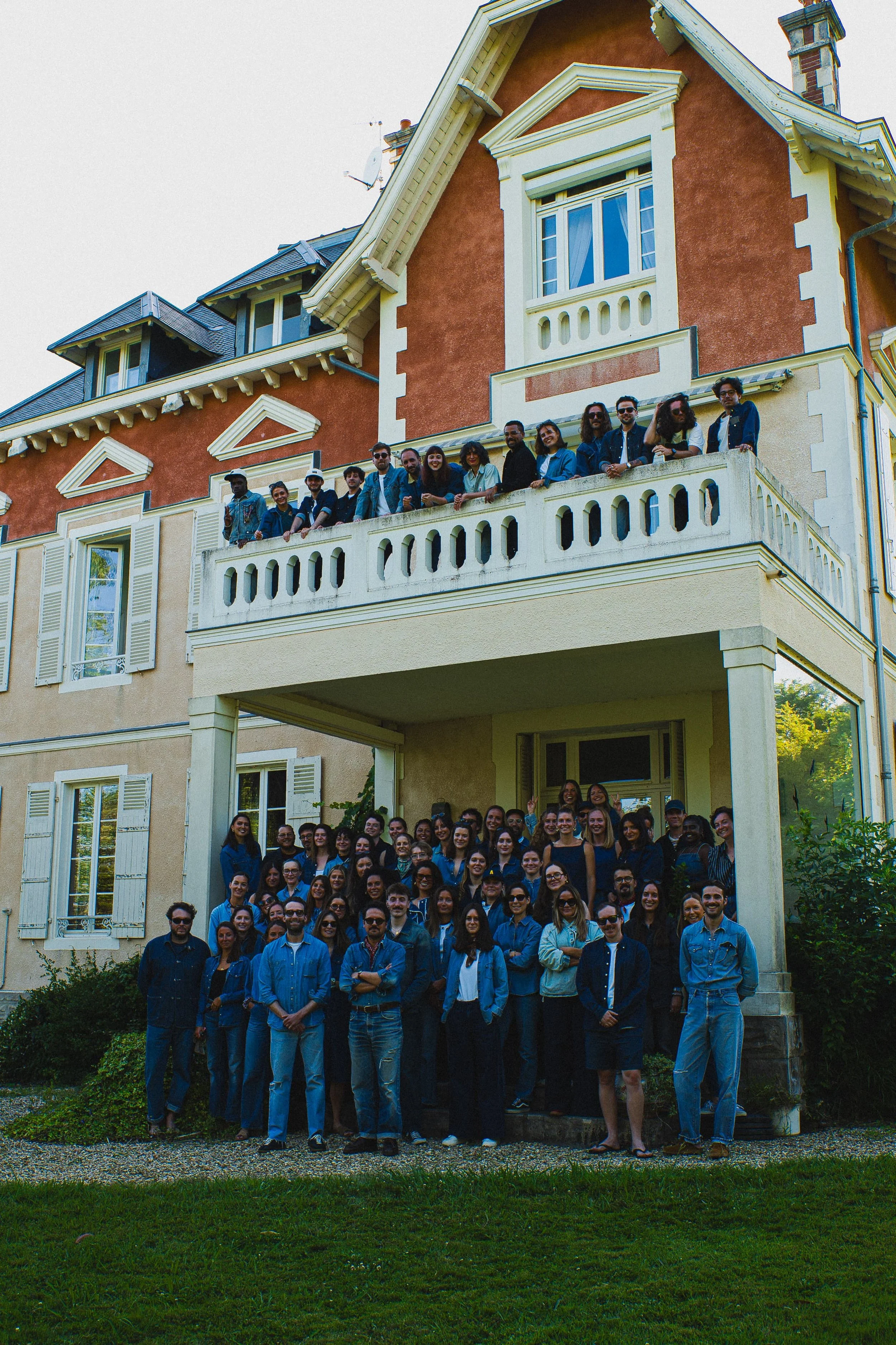 Groupe de personnes posant devant une maison de style classique avec des volets en bois blanc et une véranda. Il s'agit probablement d'une photo de groupe lors d'un événement ou d'une rencontre.