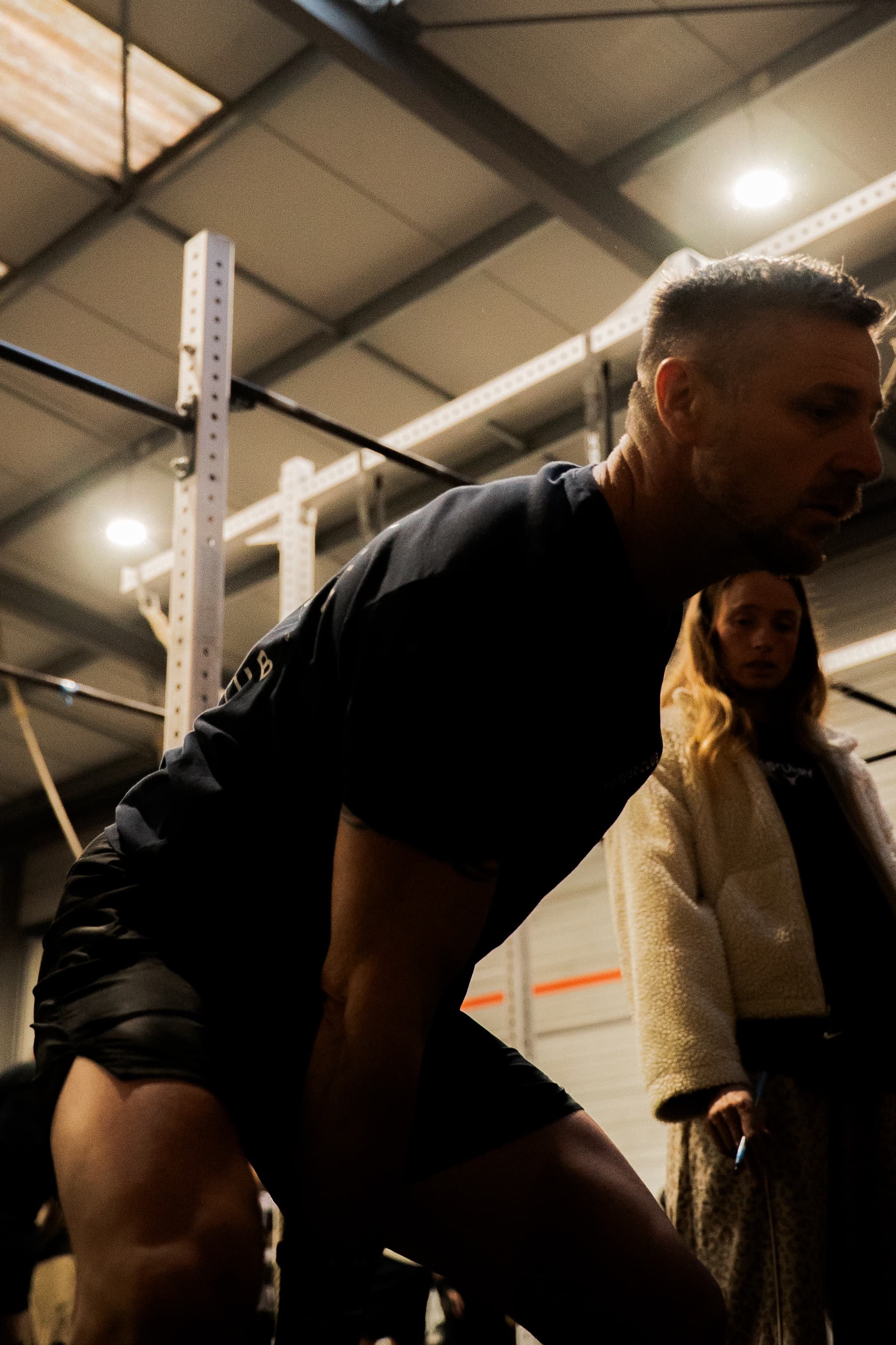 Deux personnes en train de faire de l'exercice dans une salle de sport, une personne en premier plan portant un short noir et un t-shirt noir, et une femme en arrière-plan portant un manteau beige et un pull blanc.