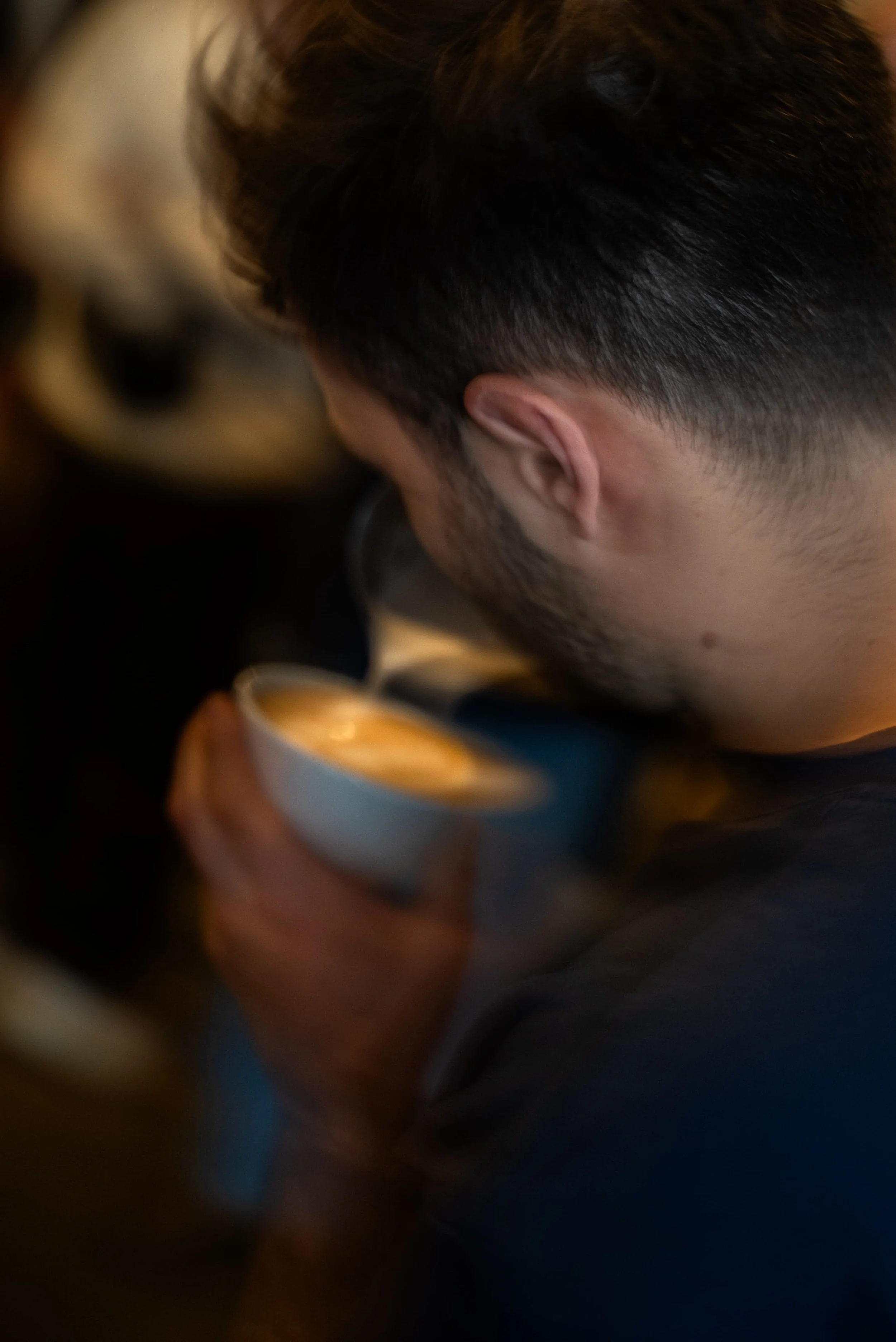 Un homme vu de dos, dégustant une boisson chaude dans une tasse, avec une chatte blanche en arrière-plan.
