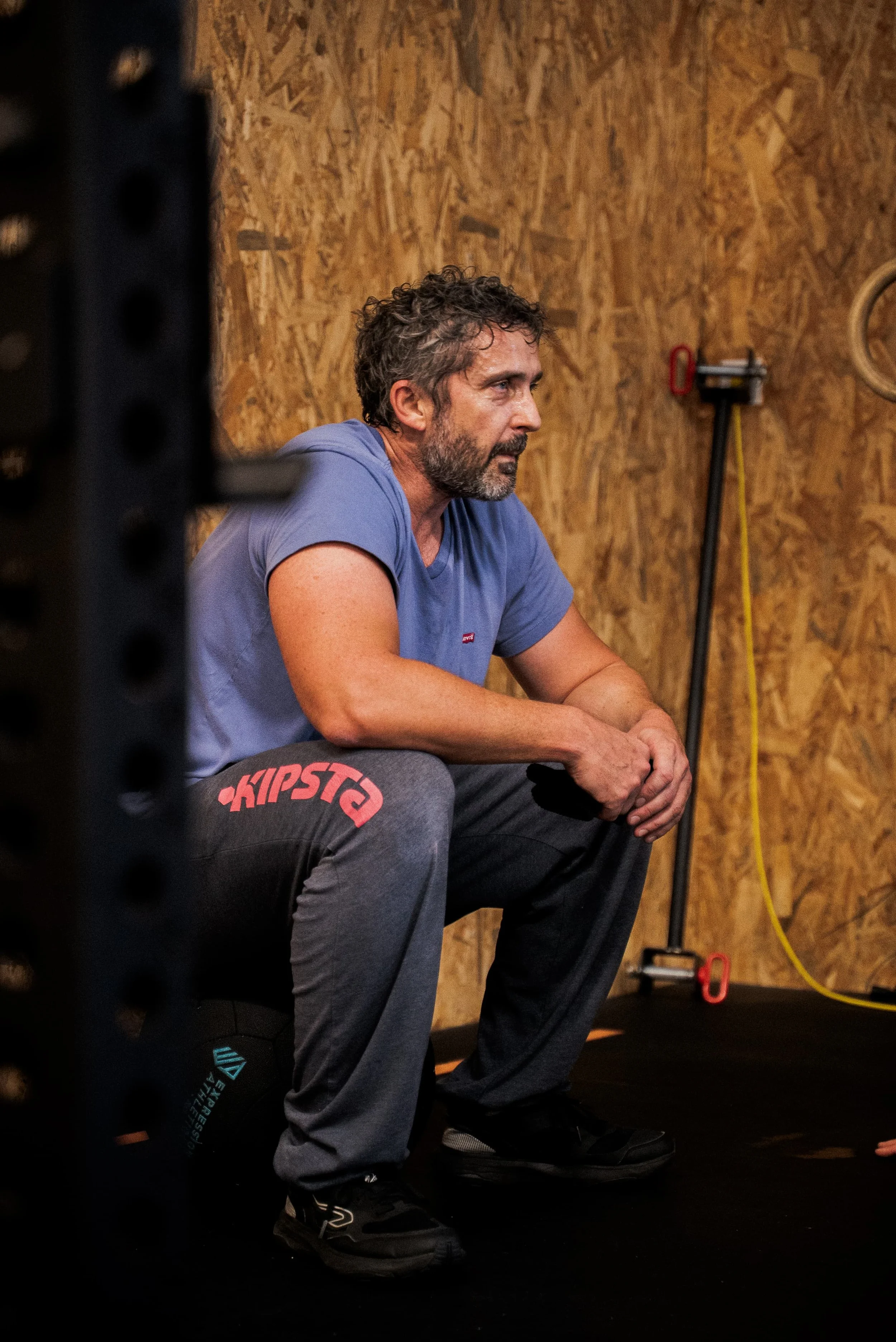 Un homme assis dans une salle de sport, portant un t-shirt bleu et un pantalon gris, semble réfléchir ou se reposer, avec un mur en bois en arrière-plan et du matériel d'entraînement à proximité.