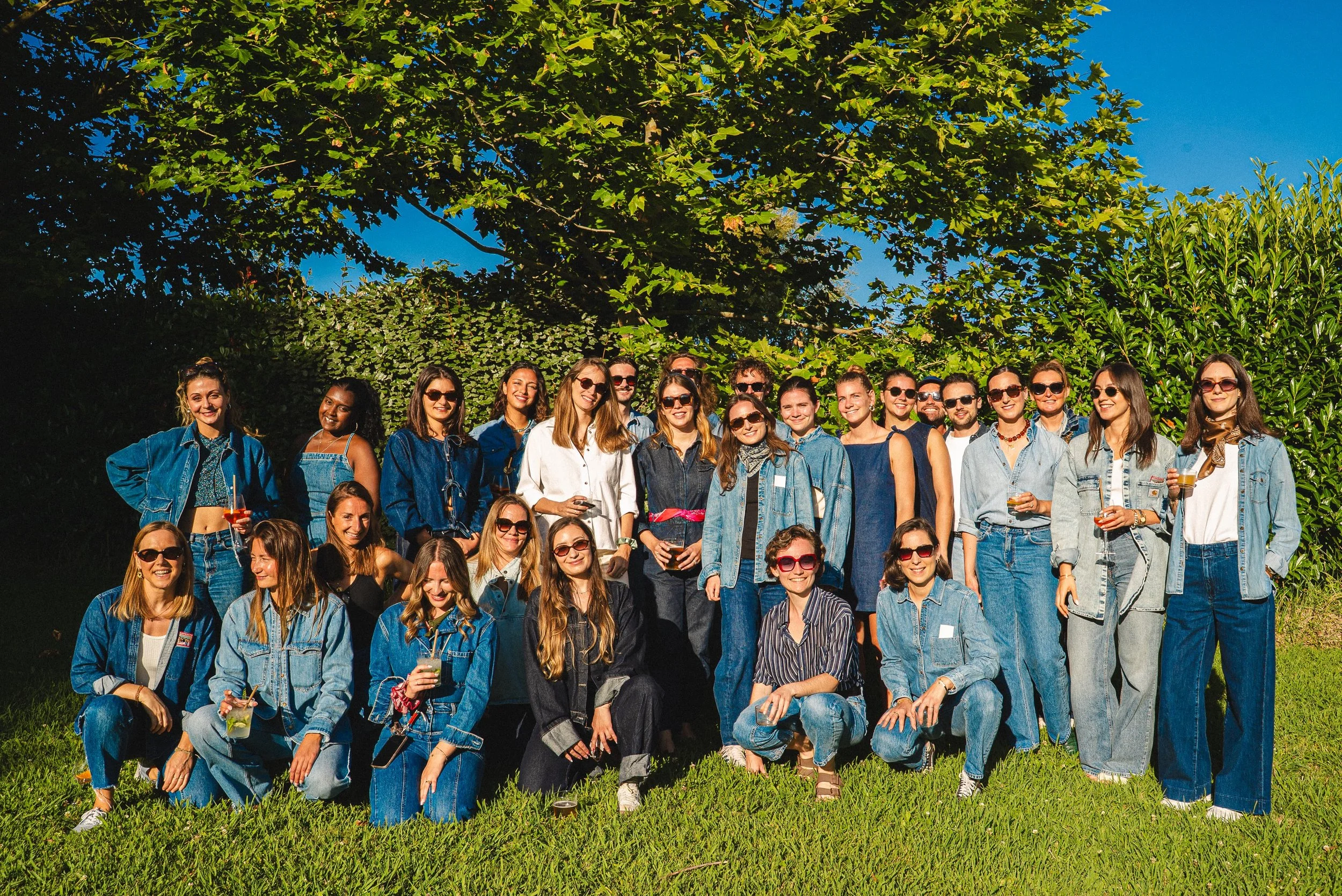 Un groupe de personnes posant pour une photo en plein air sous un grand arbre, certaines tenant des boissons, toutes souriantes.