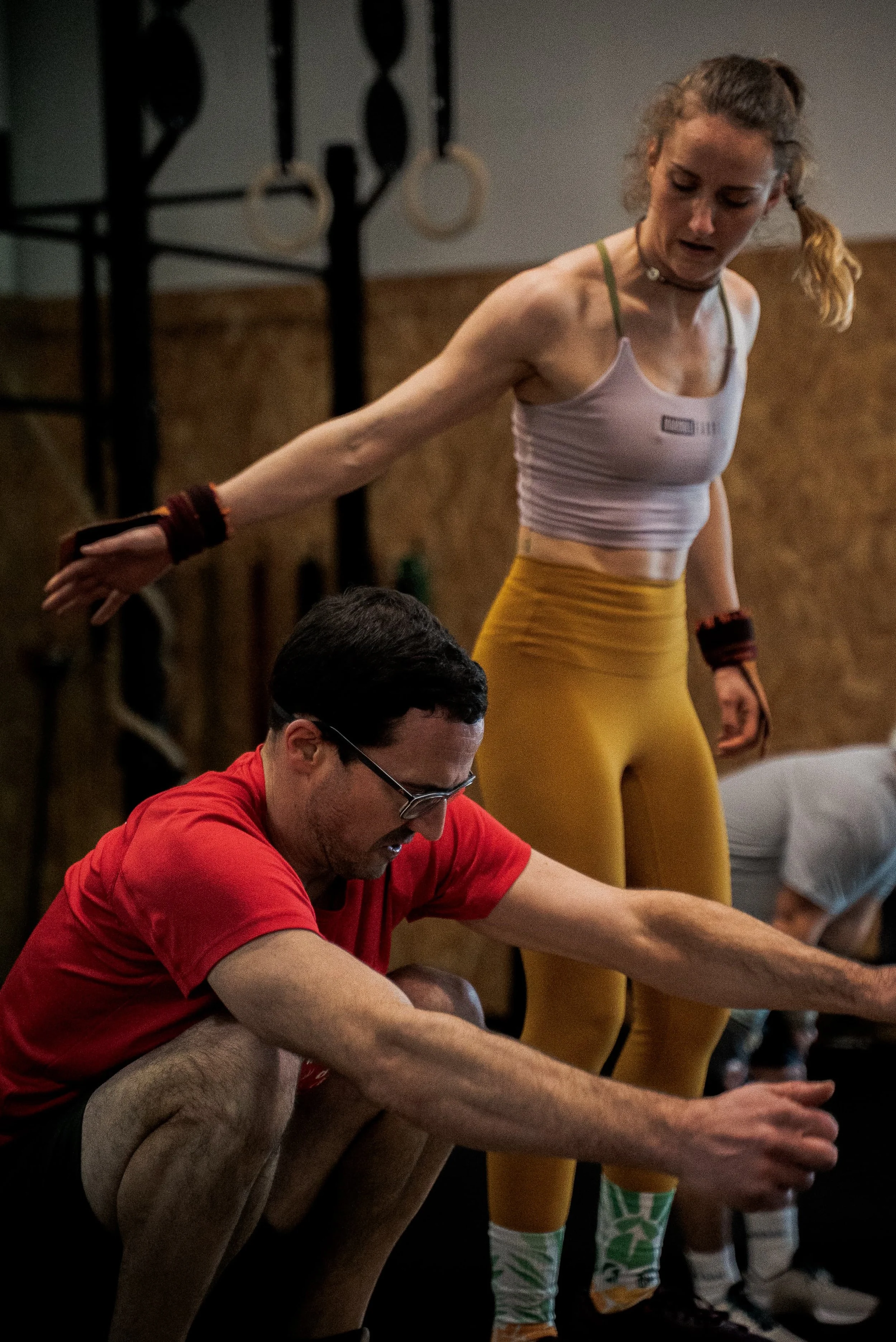 Une femme en tenue de sport, avec des leggings jaunes et un haut gris, est en train de faire des exercices sous la supervision d'un homme portant un t-shirt rouge.