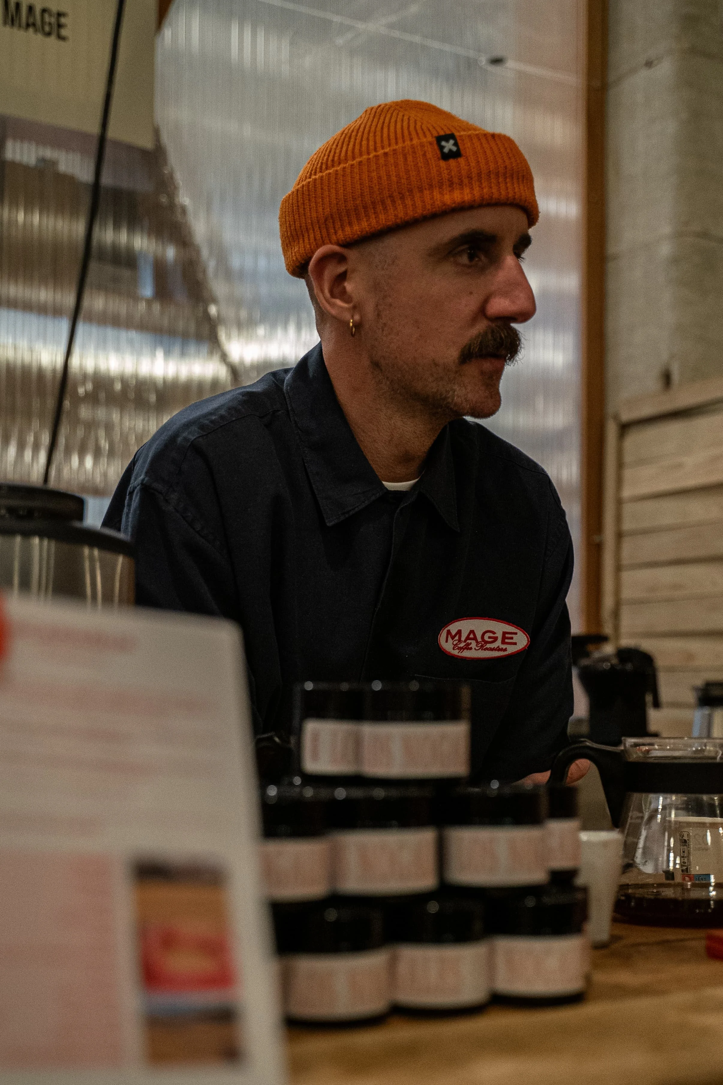 Un homme portant un bonnet orange et un uniforme de travail en train de travailler dans un café, assis derrière un comptoir en bois.