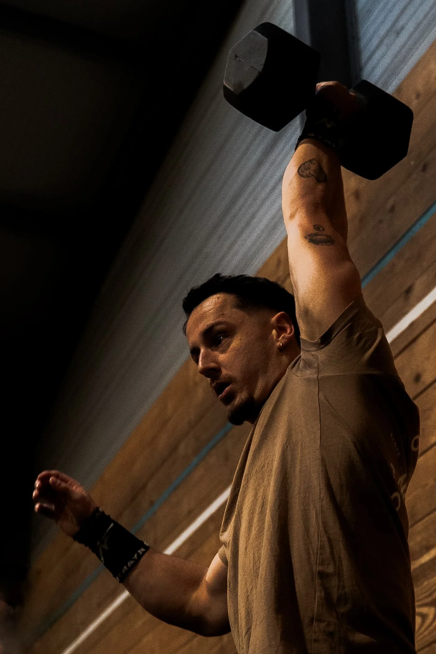 Un homme avec des tatouages sur le bras, portant un t-shirt beige, soulève un haltère dans une salle de sport, avec un mur en bois en arrière-plan.