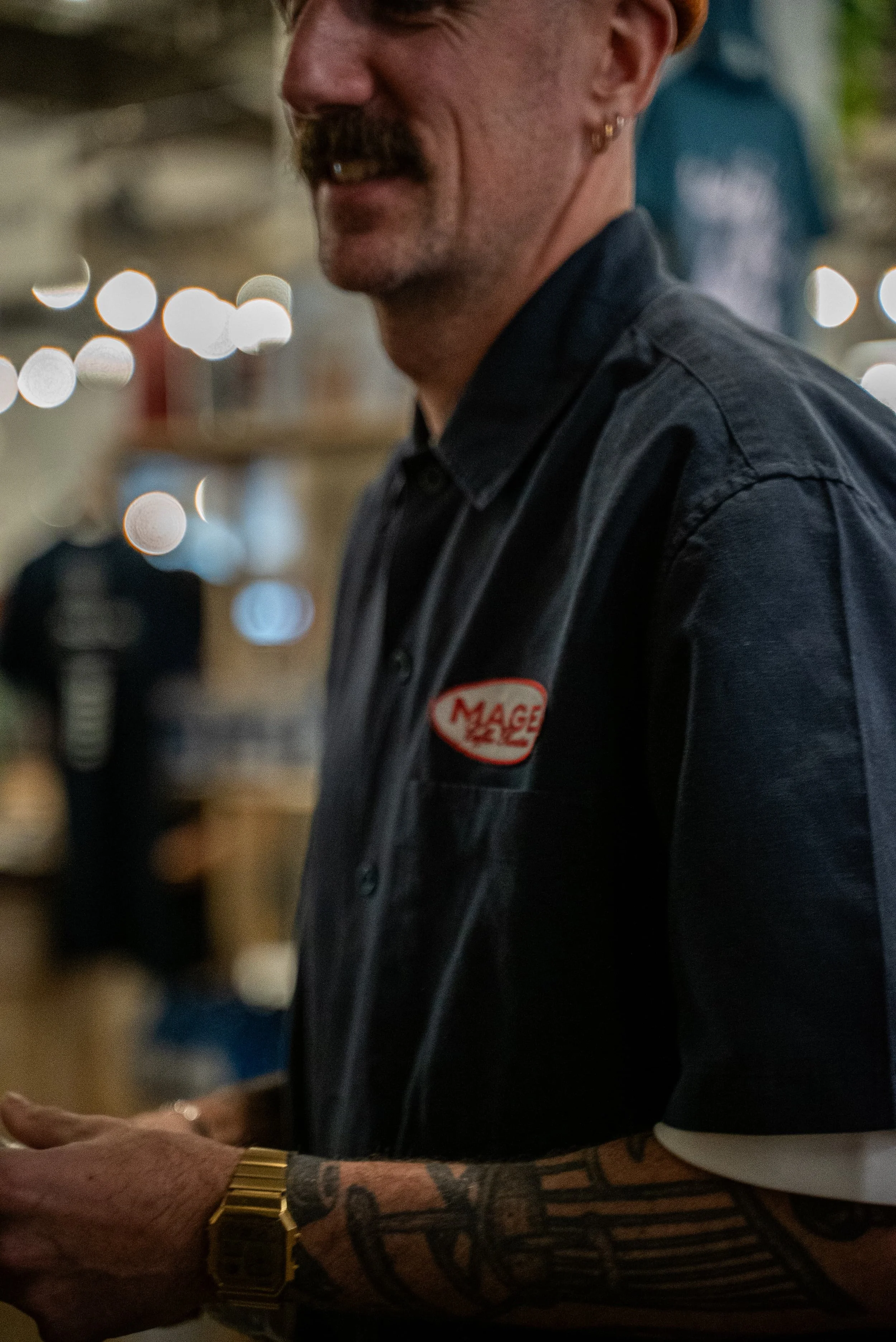 Close-up d'un homme portant une chemise en jean avec une étiquette 'MAGE' et une montre en or, avec un tatouage sur le bras, dans un endroit lumineux.
