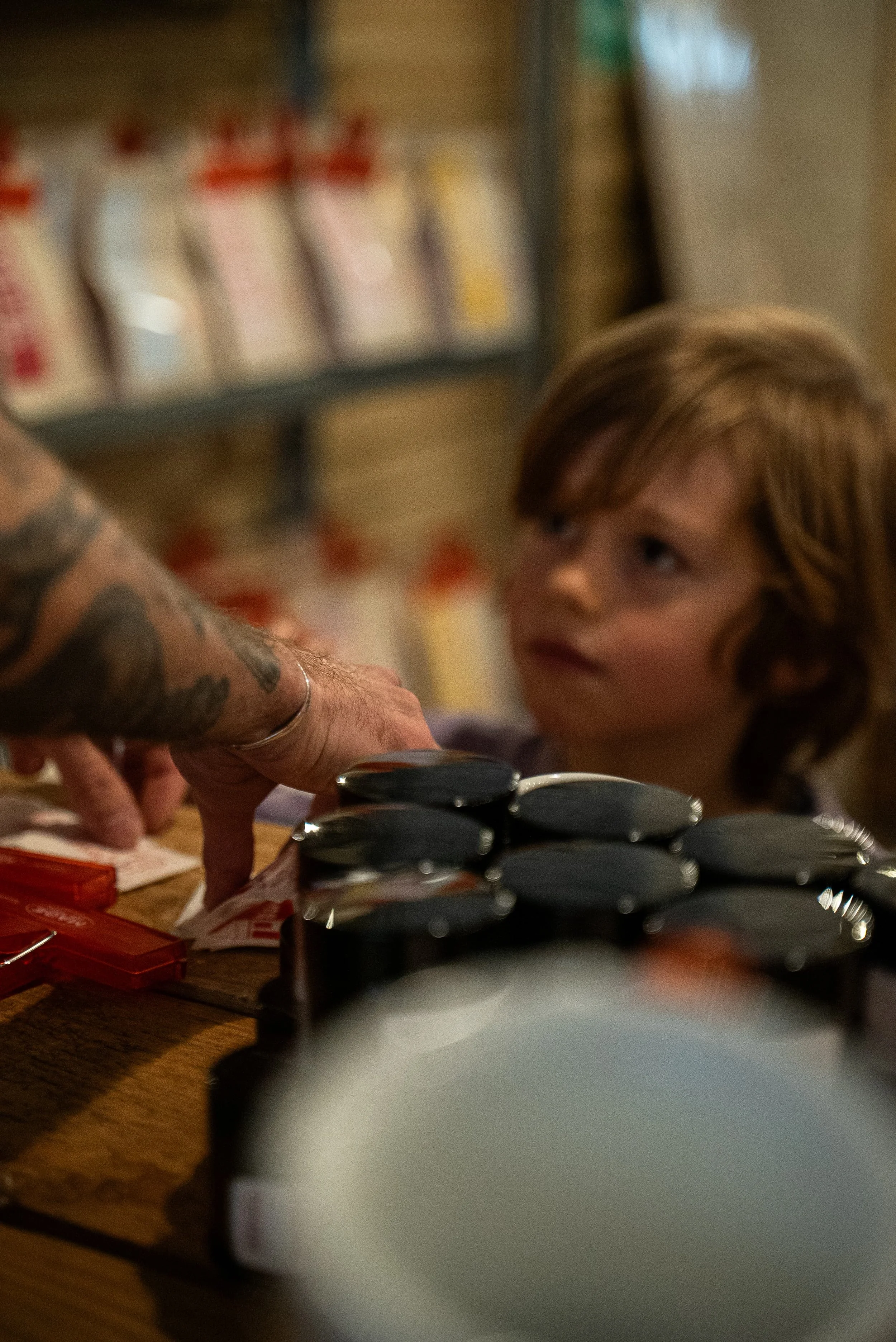 Jeune garçon blond regardant une main tatouée poser des cigarettes sur une table en bois, avec un plat à côté.