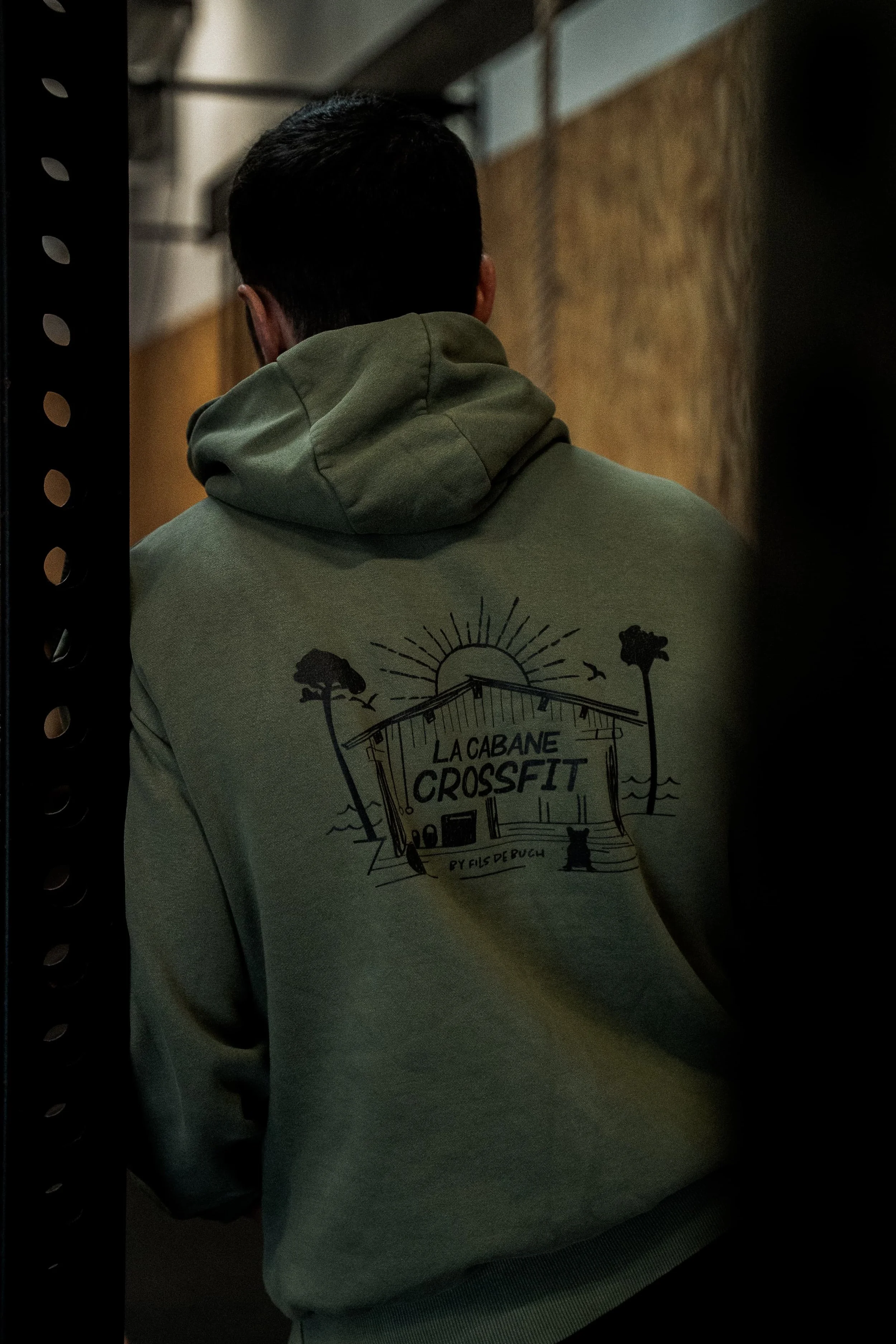 Person vue de dos portant un sweat à capuche gris avec un dessin représentant une cabane de CrossFit, des arbres et un soleil, avec le texte La Cabane CrossFit.