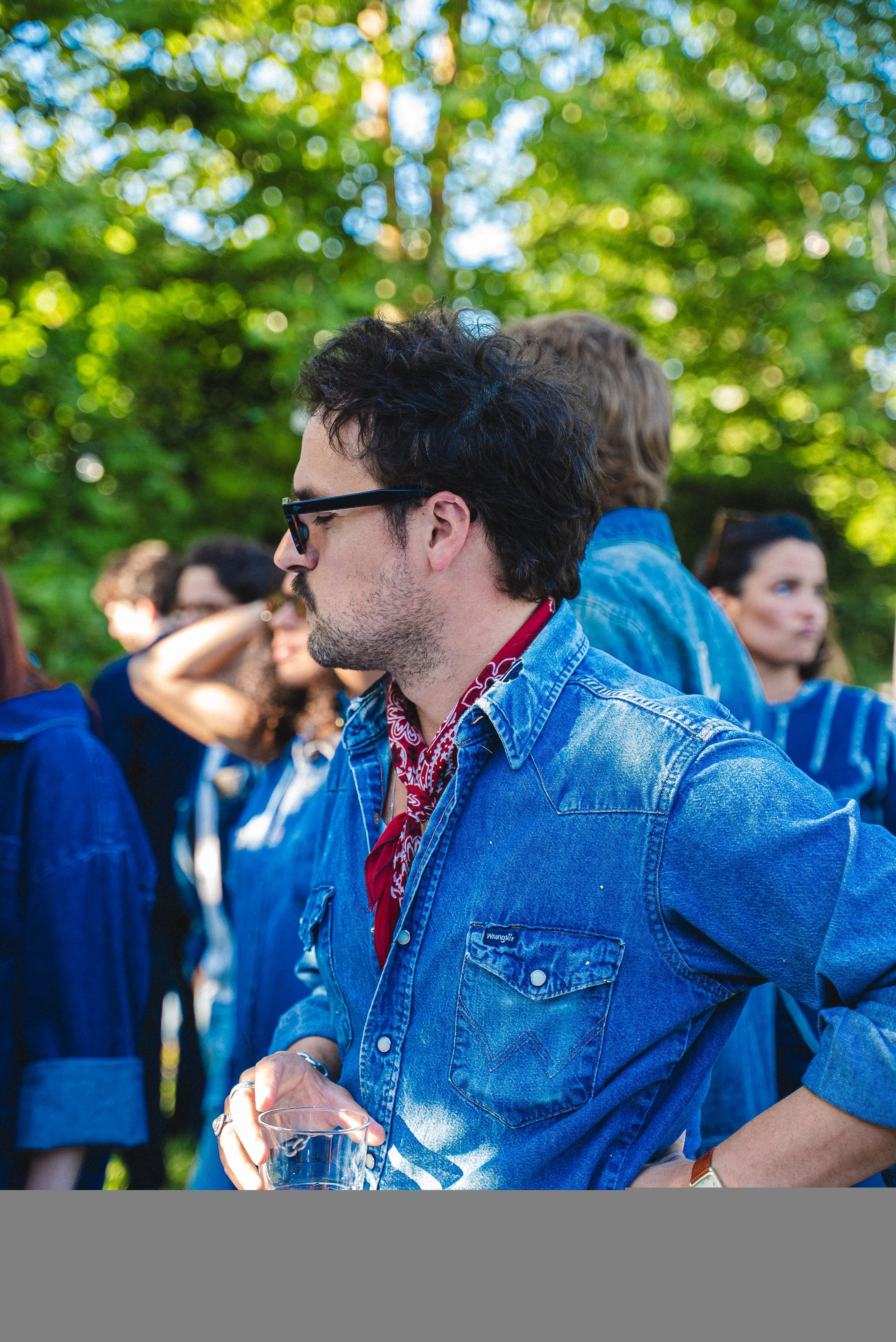 Un homme portant des lunettes de soleil, une chemise en jean et un bandana rouge, tenant un verre d'eau, lors d'un rassemblement en plein air entouré de plusieurs autres personnes, avec une ambiance décontractée et ensoleillée, dans un environnement 