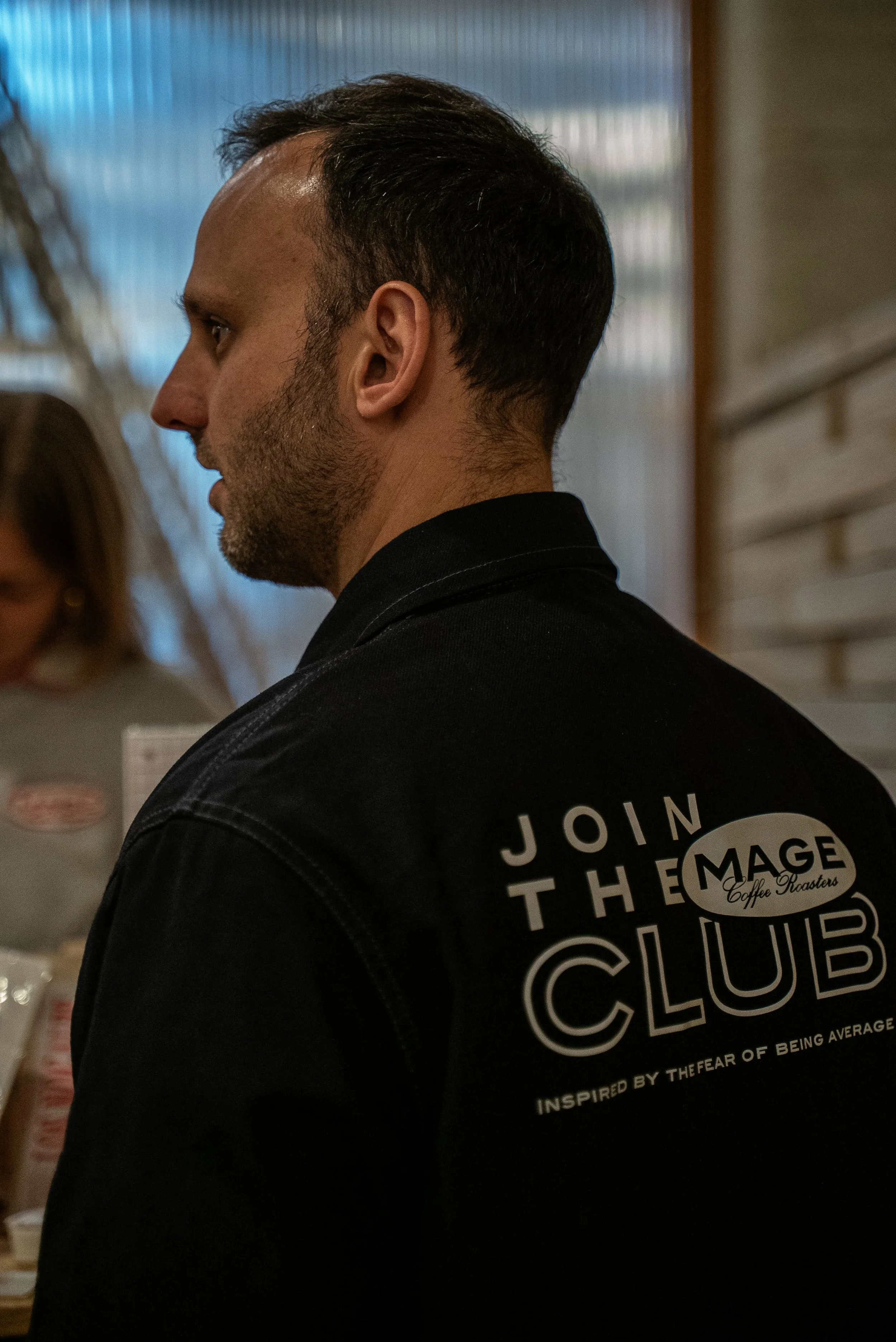 Profil d'un homme avec une coupe de cheveux courte sur les côtés et plus longue sur le dessus, portant un t-shirt noir avec un logo ou un texte en blanc.