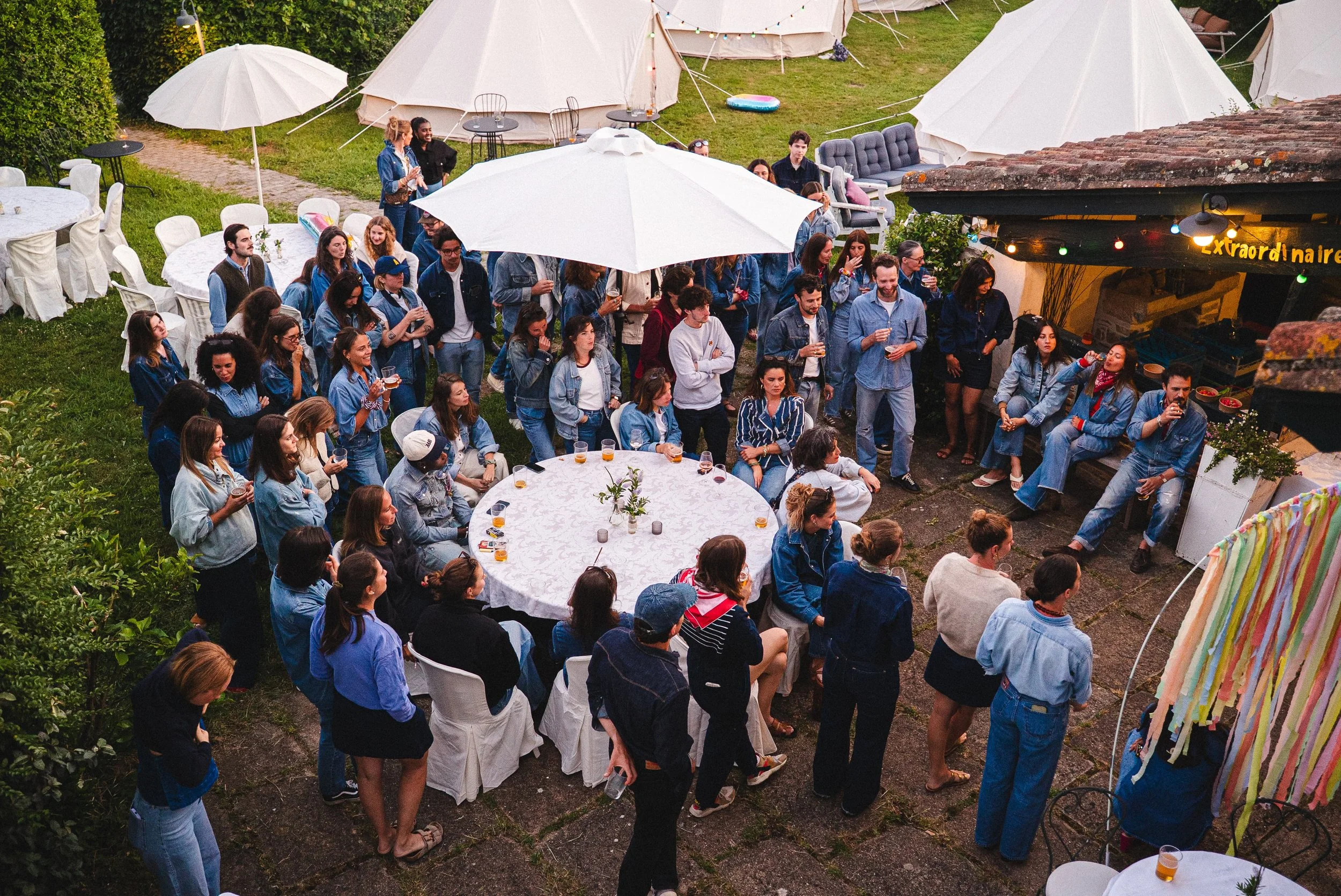 Un groupe de personnes lors d'une fête ou d'une réunion en extérieur, sous des parasols en soirée, avec des tentes en toile de camping en arrière-plan.