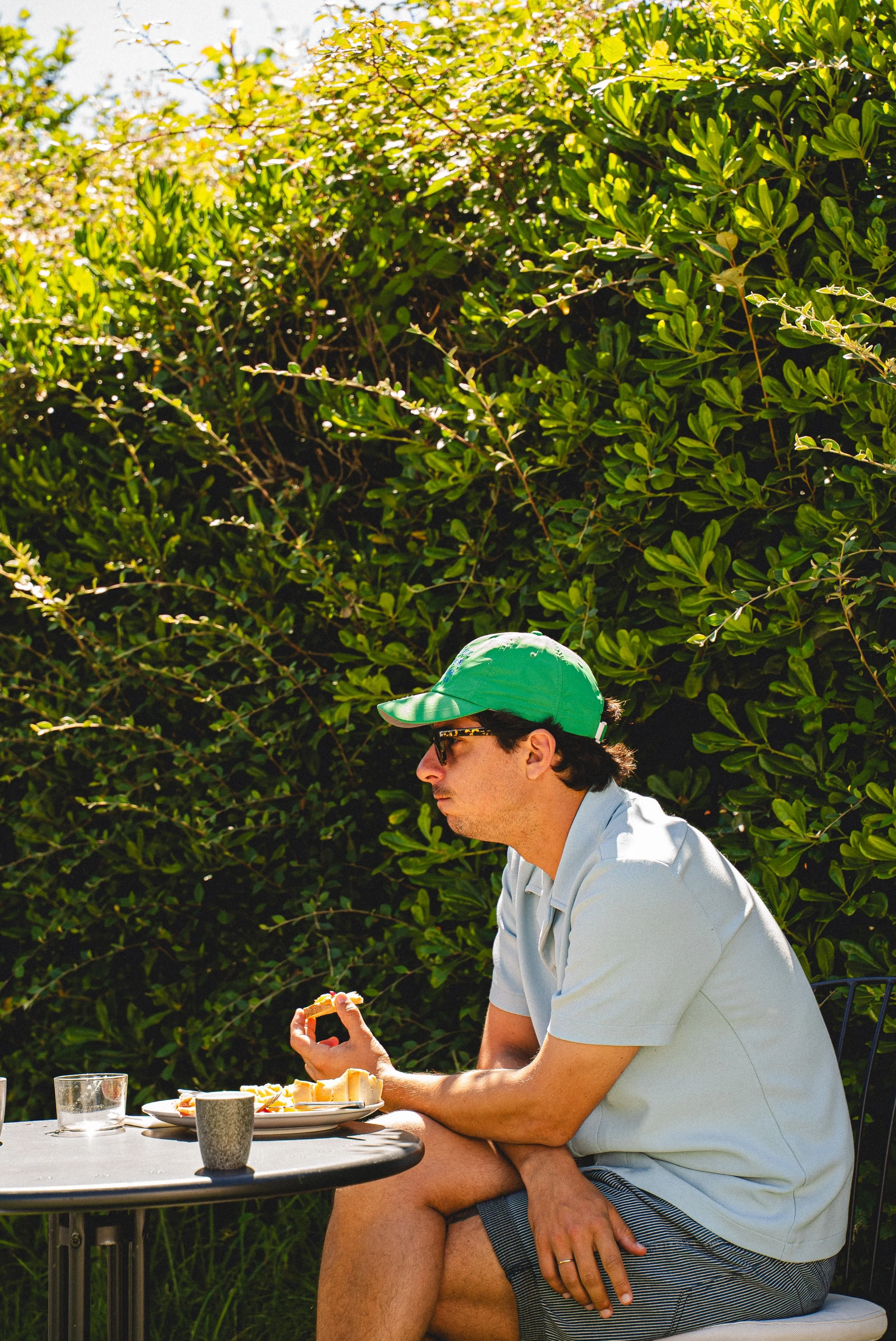 Un homme portant un chapeau vert, des lunettes de soleil, une chemisette blanche, et des shorts à rayures, est assis à une table en extérieur, tenant une tranche de gâteau dans la main, avec un plateau de nourriture, des verres, et un pot à côté, dev