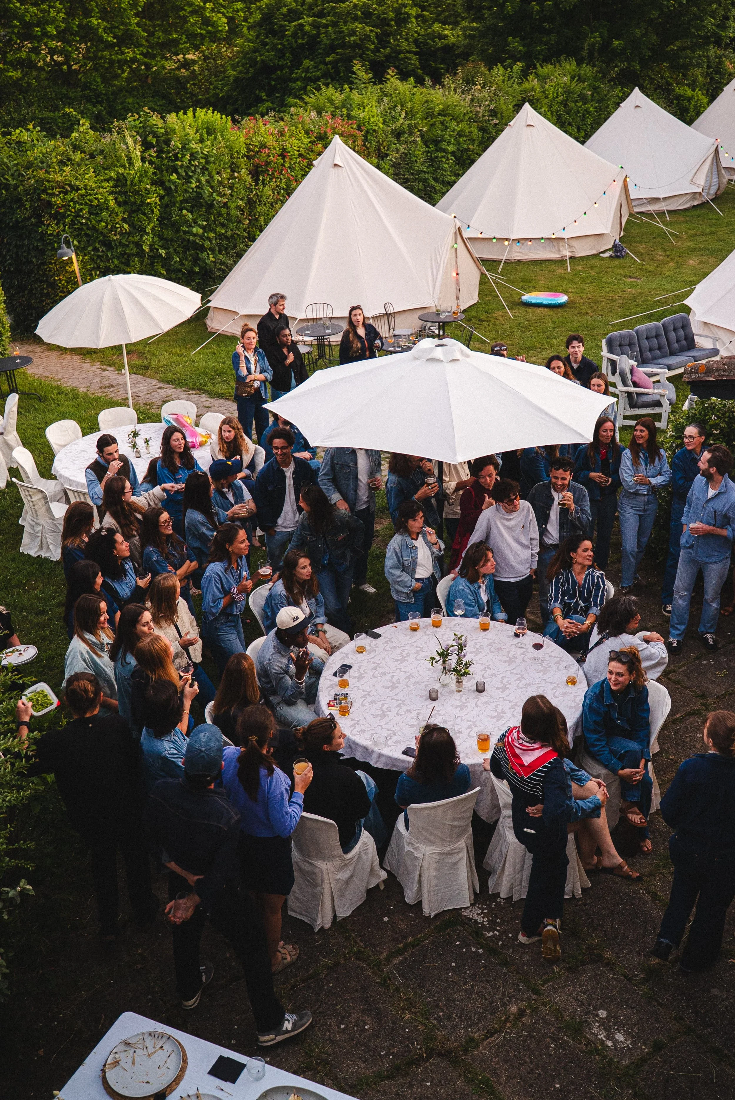Une fête en plein air avec de nombreux invités réunis autour de tables rondes sous des parasols blancs, dans un jardin avec des tentes blanches en toile de fond, décorées de guirlandes lumineuses.