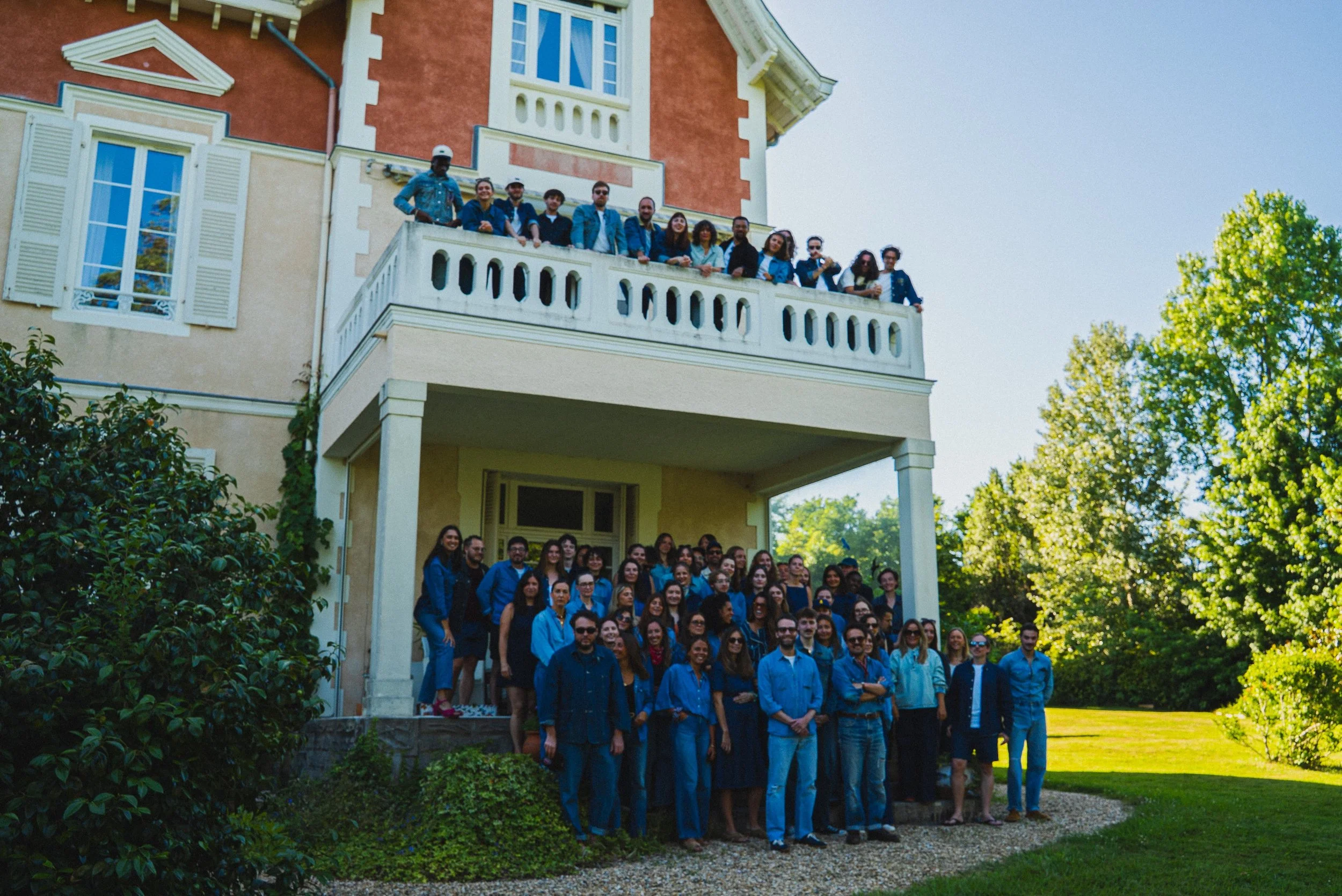 Un groupe de personnes posant devant une maison avec un balcon, sous un ciel clair, entouré d'arbres verts, lors d'une journée ensoleillée.