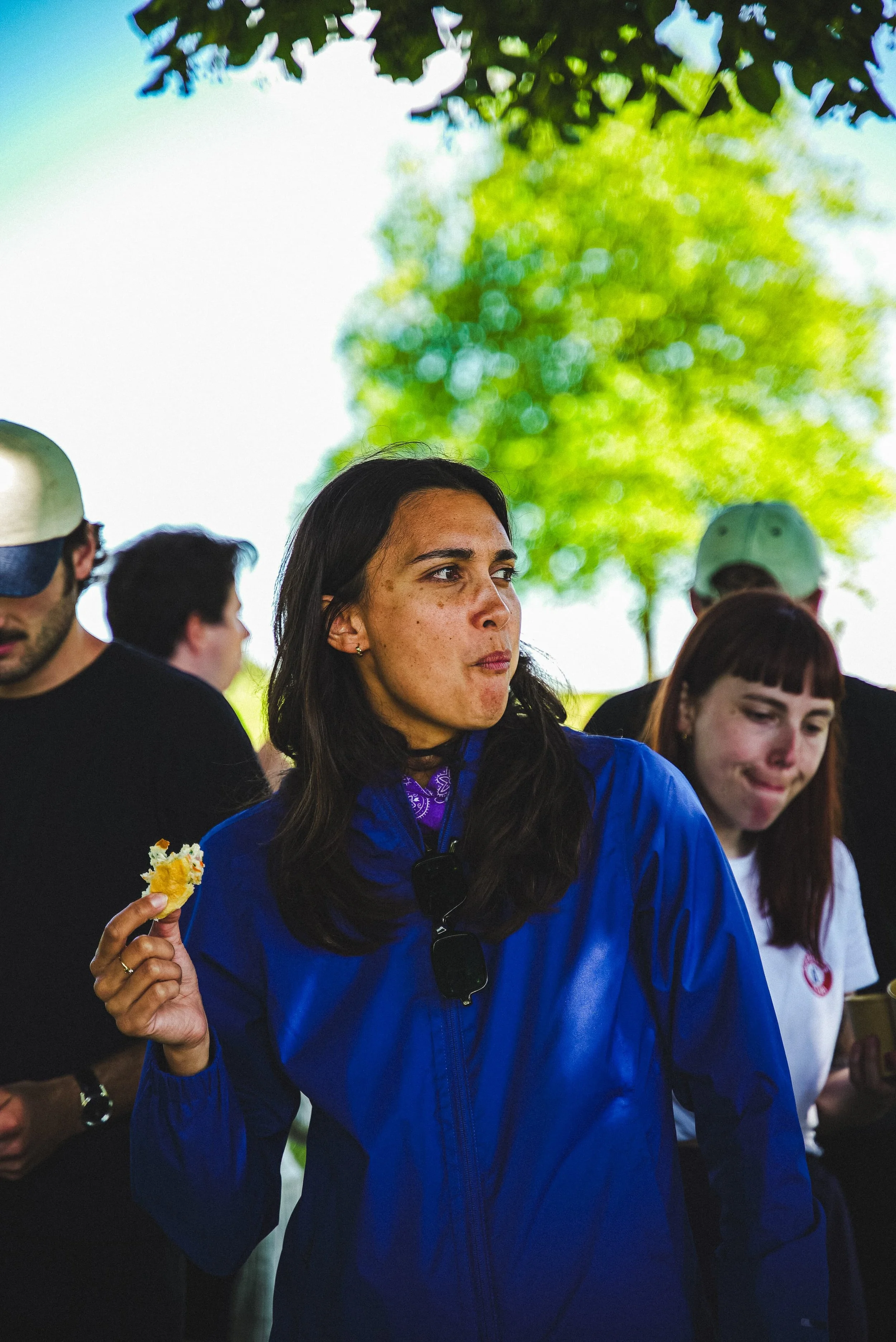 Une femme en veste bleue tenant un morceau de tarte ou de gâteau dans la main, entourée d'autres personnes dans un espace extérieur avec des arbres en arrière-plan.