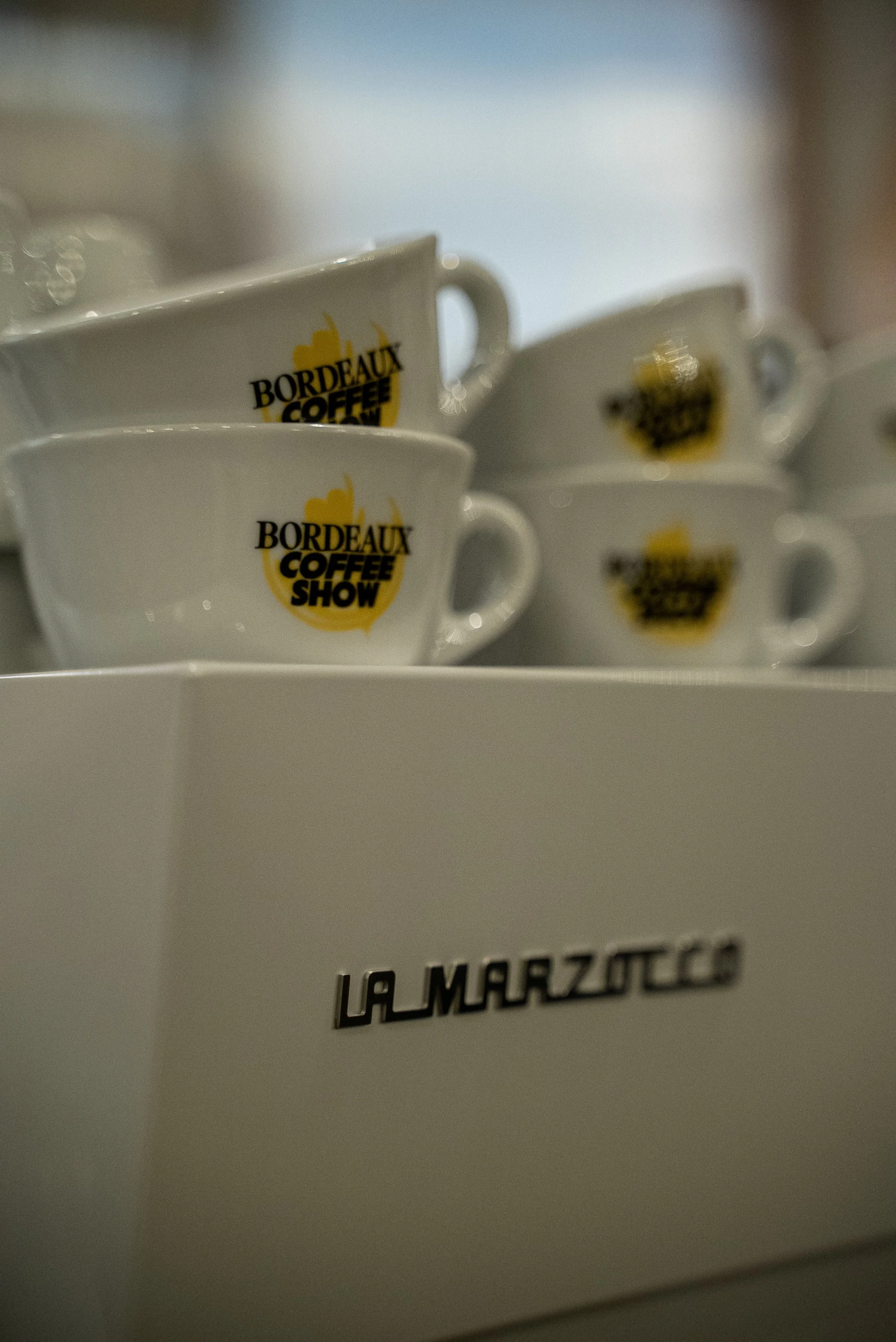 Tasses blanches avec le logo 'Bordeaux Coffee Show' en jaune et noir, empilées sur une surface blanche, avec un fond flou. 