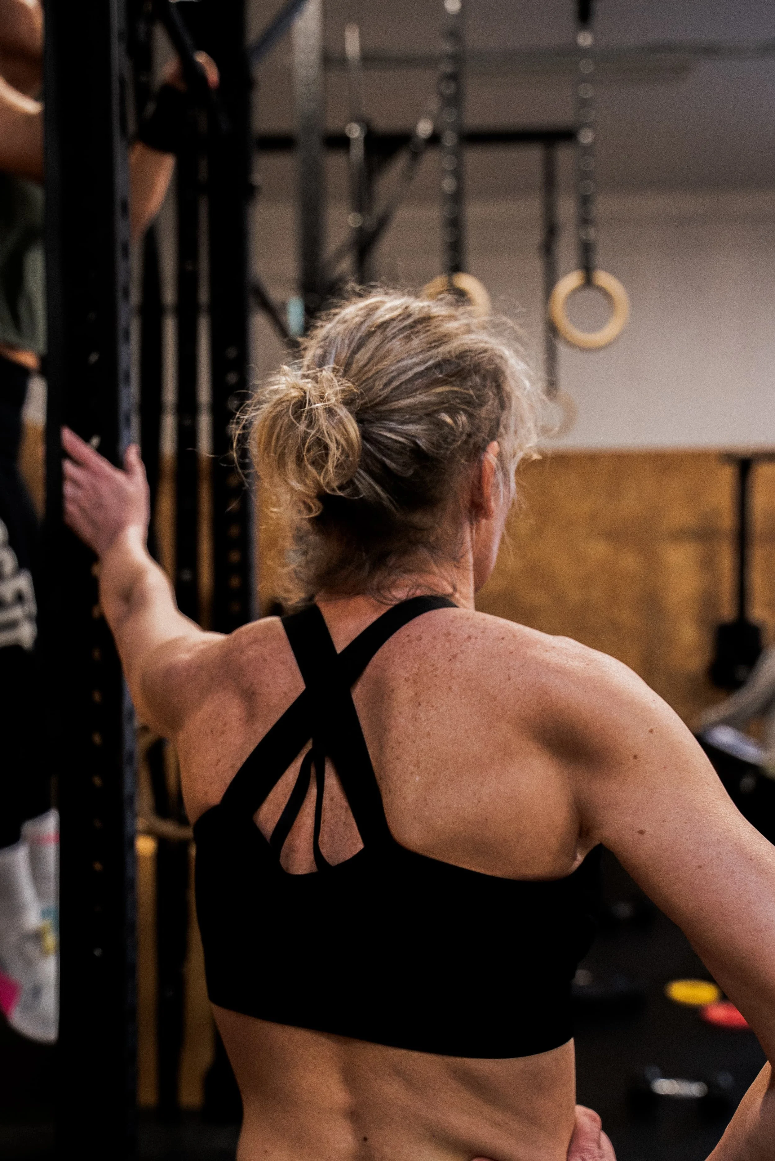 Femme musclée portant un vêtement de sport noir, vue de dos, dans une salle de sport avec des équipements d'entraînement.