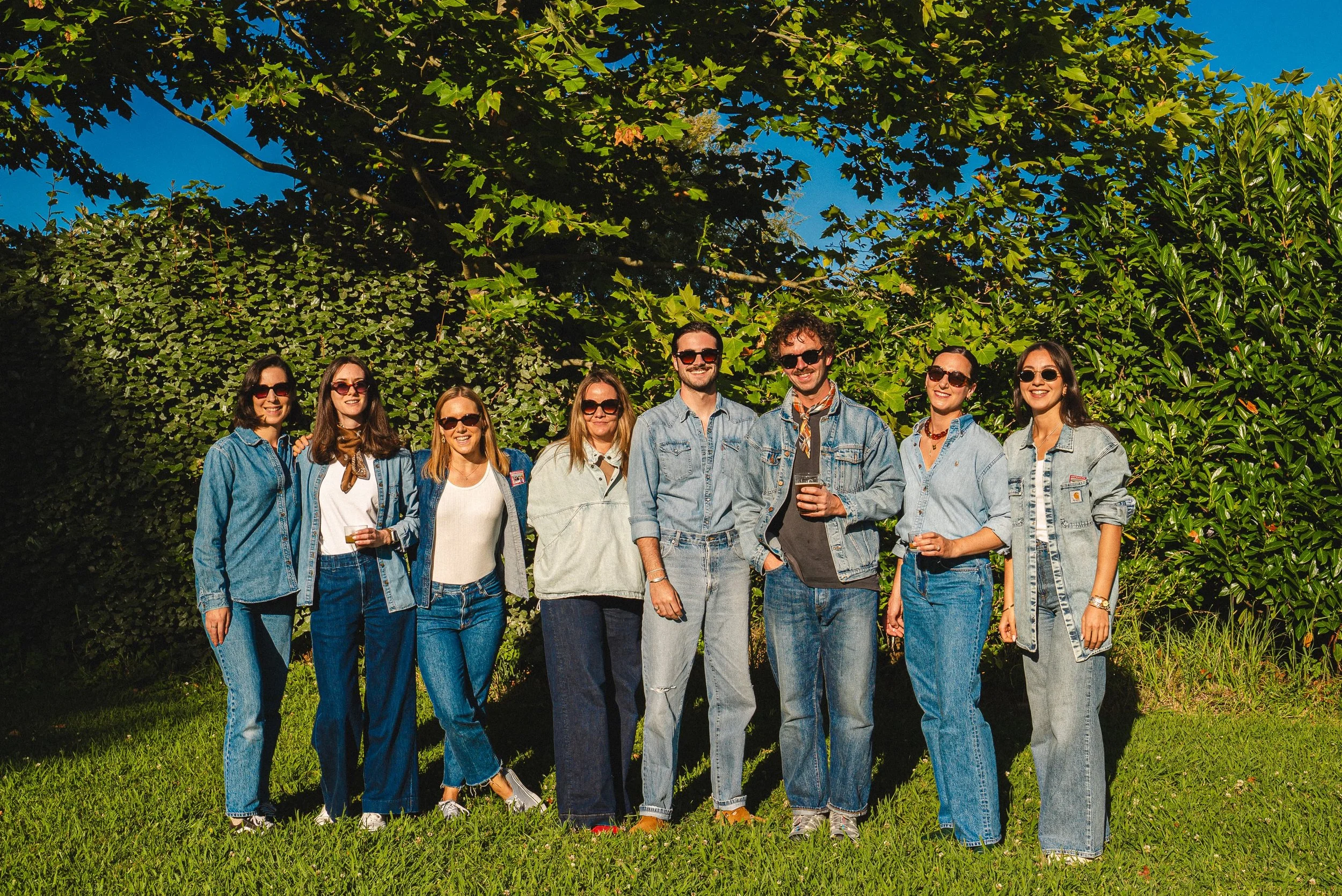 Un groupe de neuf jeunes adultes debout sur l'herbe en plein air, portant des vêtements en denim et des lunettes de soleil, souriants sous un ciel bleu, entourés de végétation verte.