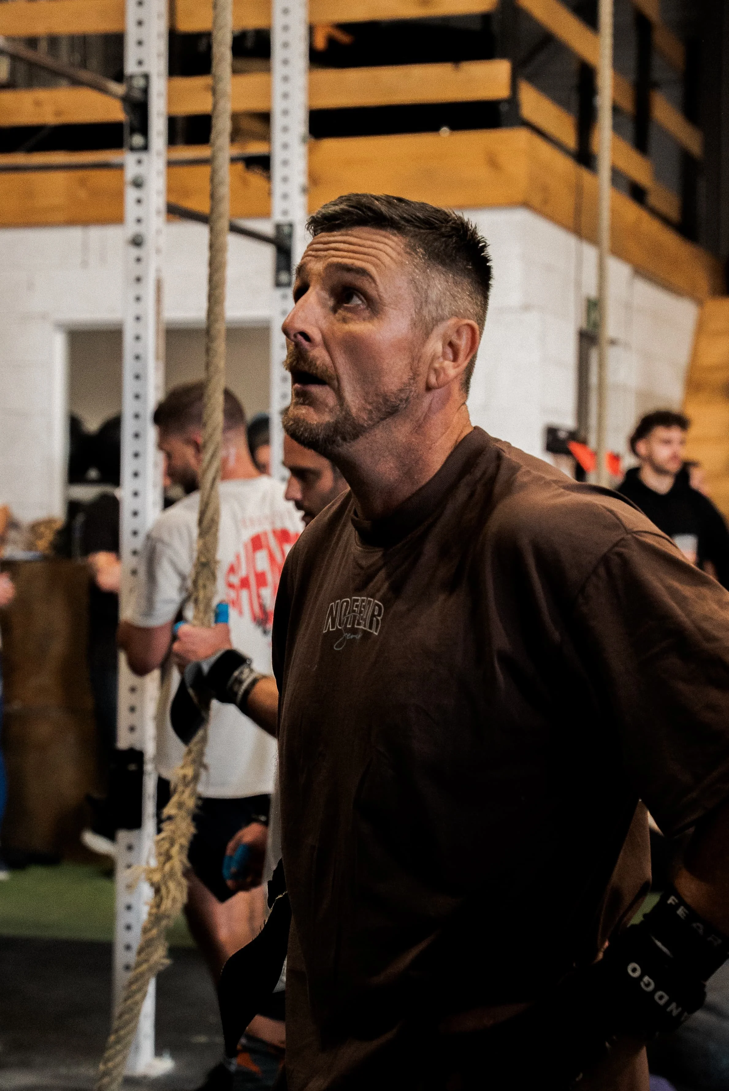 Un homme avec une barbe court dans une salle de sport, portant une veste marron et des gants de boxe, entouré d'autres personnes qui s'entraînent.