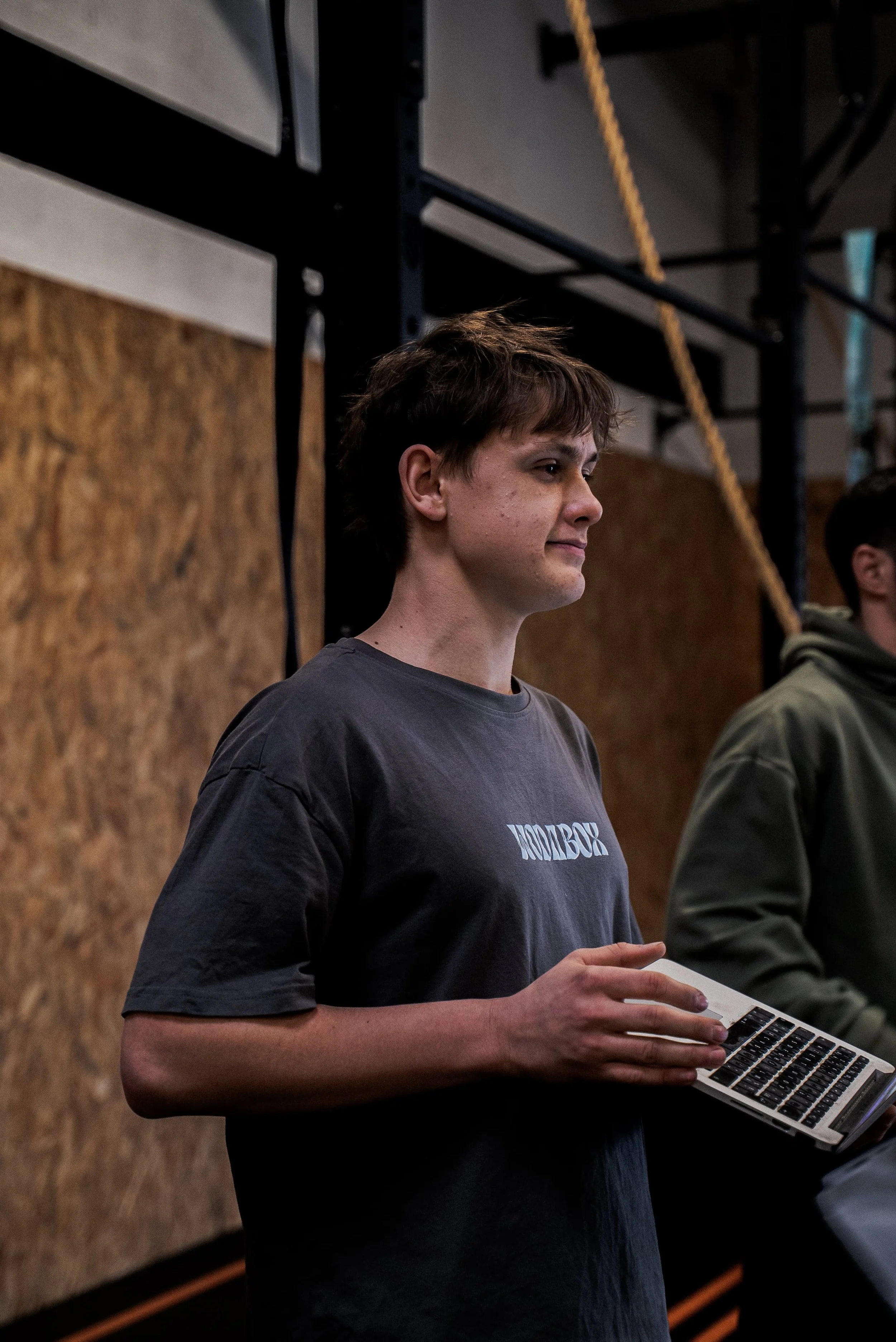 Jeune homme avec des cheveux bruns courts, portant un t-shirt gris, tenant un clavier d'ordinateur portable dans un environnement intérieur avec un mur en bois en arrière-plan.