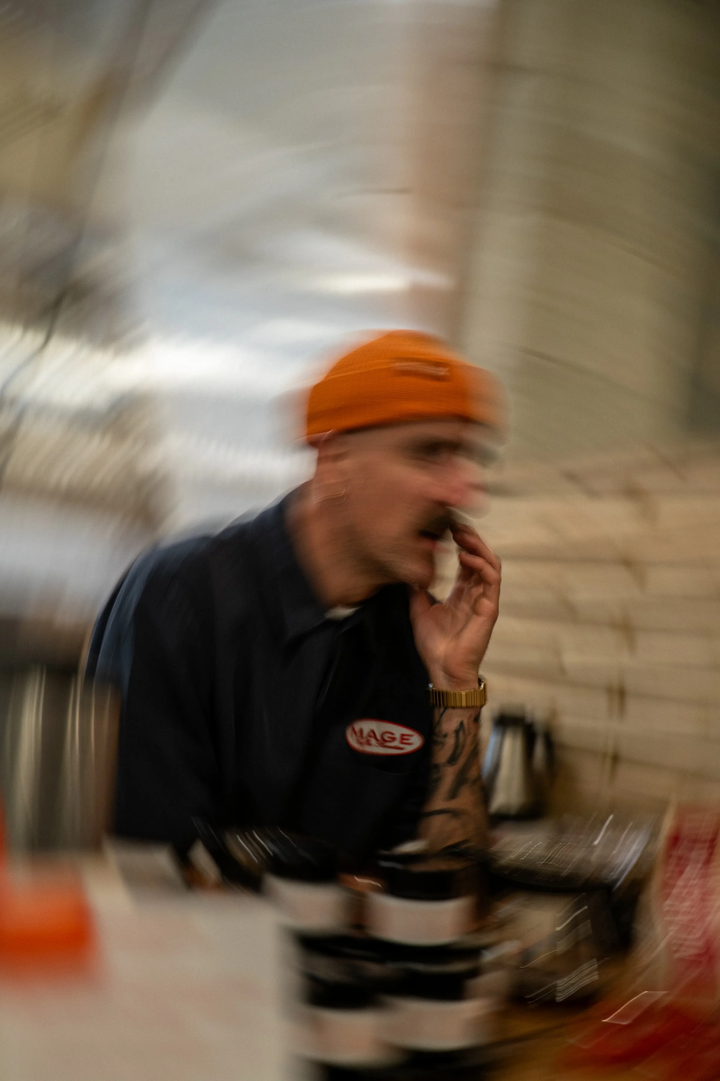 Une personne portant un bonnet orange, assise à une table, tenant un téléphone à l'oreille, avec des plats et des objets sur la table dans un décor intérieur.