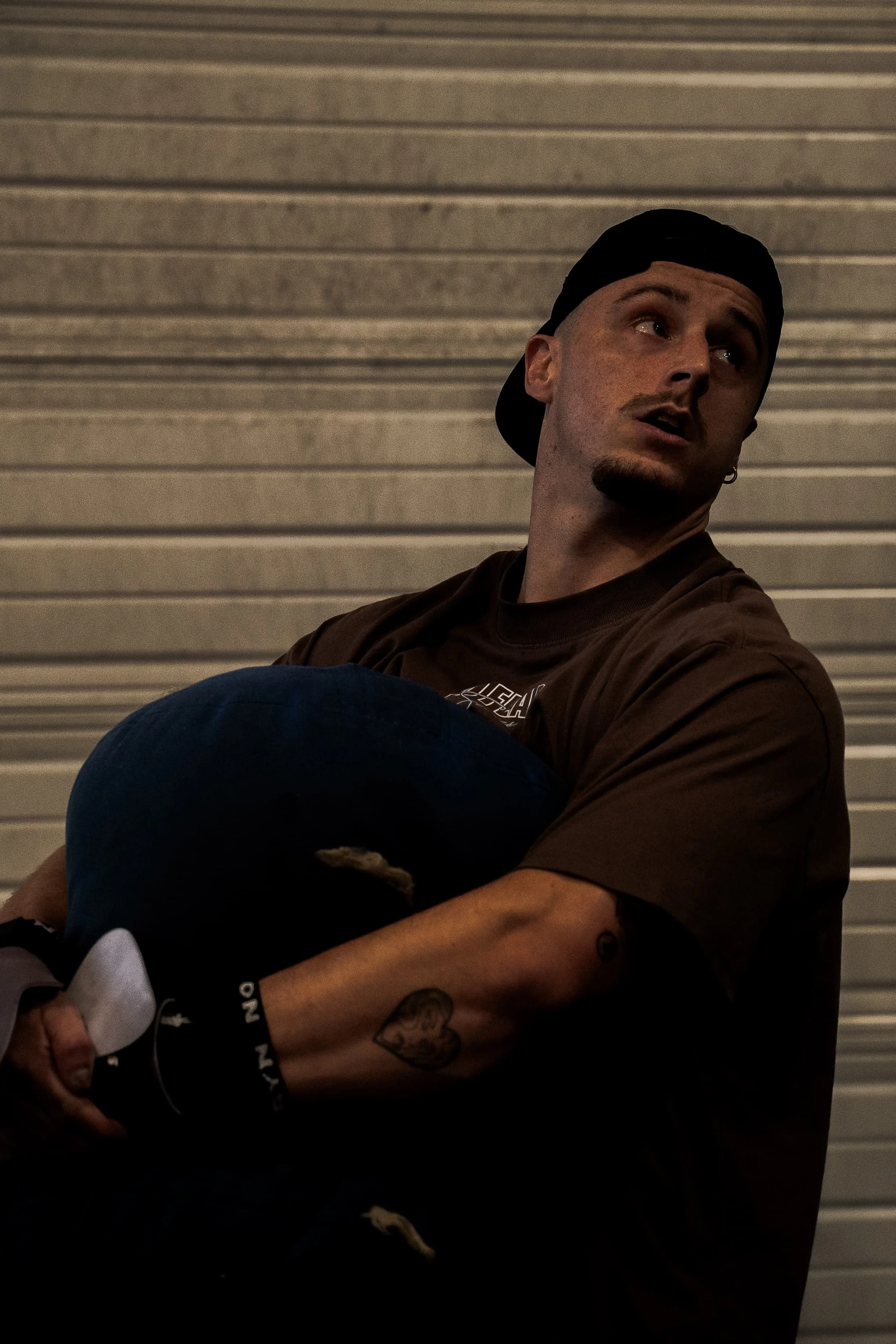Un jeune homme portant une casquette noire, un t-shirt marron et une montre noire, assis contre un mur en métal, regardant vers la droite, avec un tatouage de visage sur le bras.