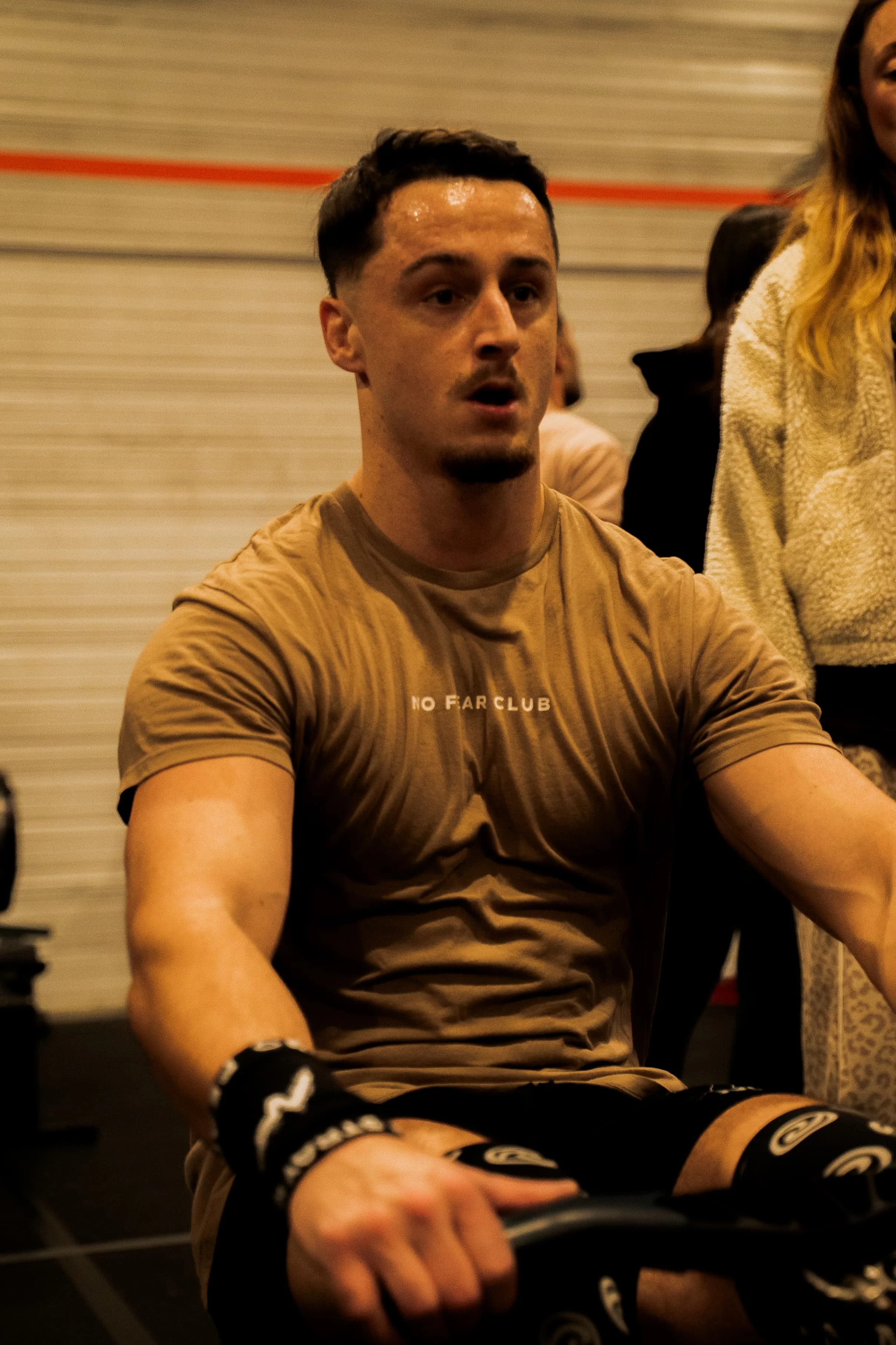 Un homme en sweat marron assis, portant des gants de boxe, dans une salle de sport.