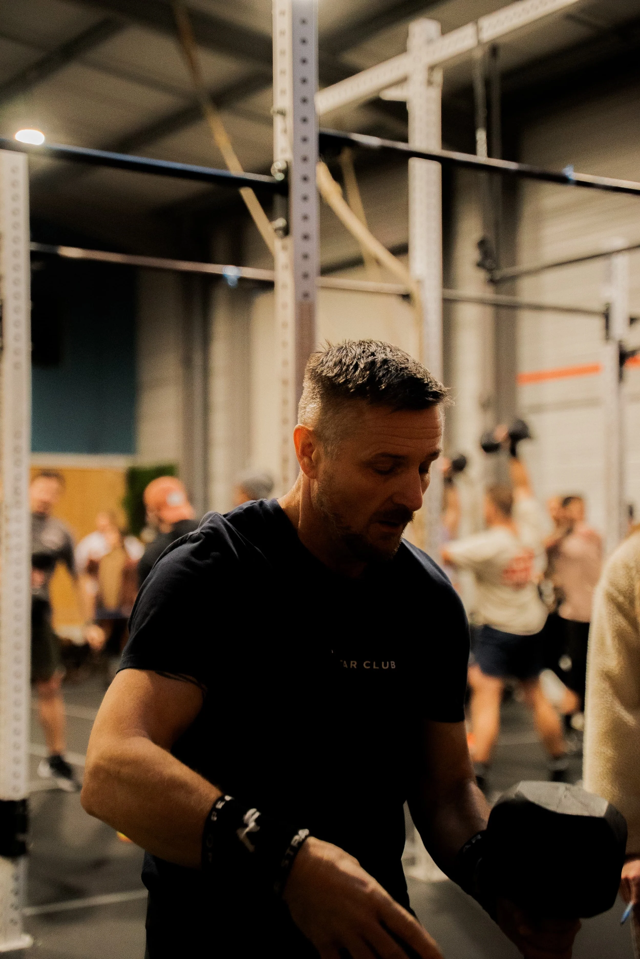 Un homme aux cheveux courts et bouclés en train de se préparer dans un gymnase, avec d'autres personnes en arrière-plan.
