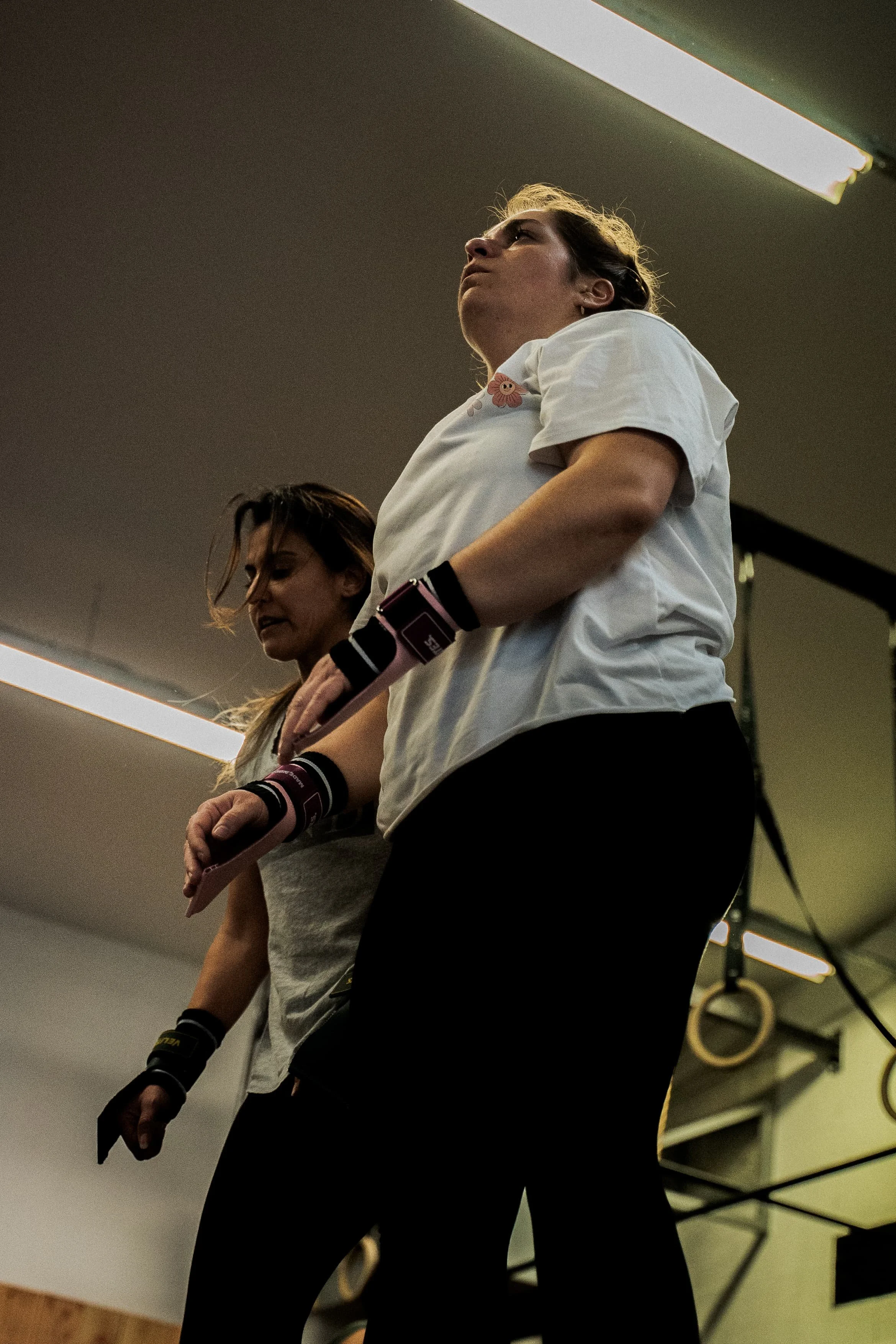 Deux femmes s'entraînent dans une salle de sport, tenant des haltères, vues de côté avec un éclairage de plafond, dans une ambiance sportive.