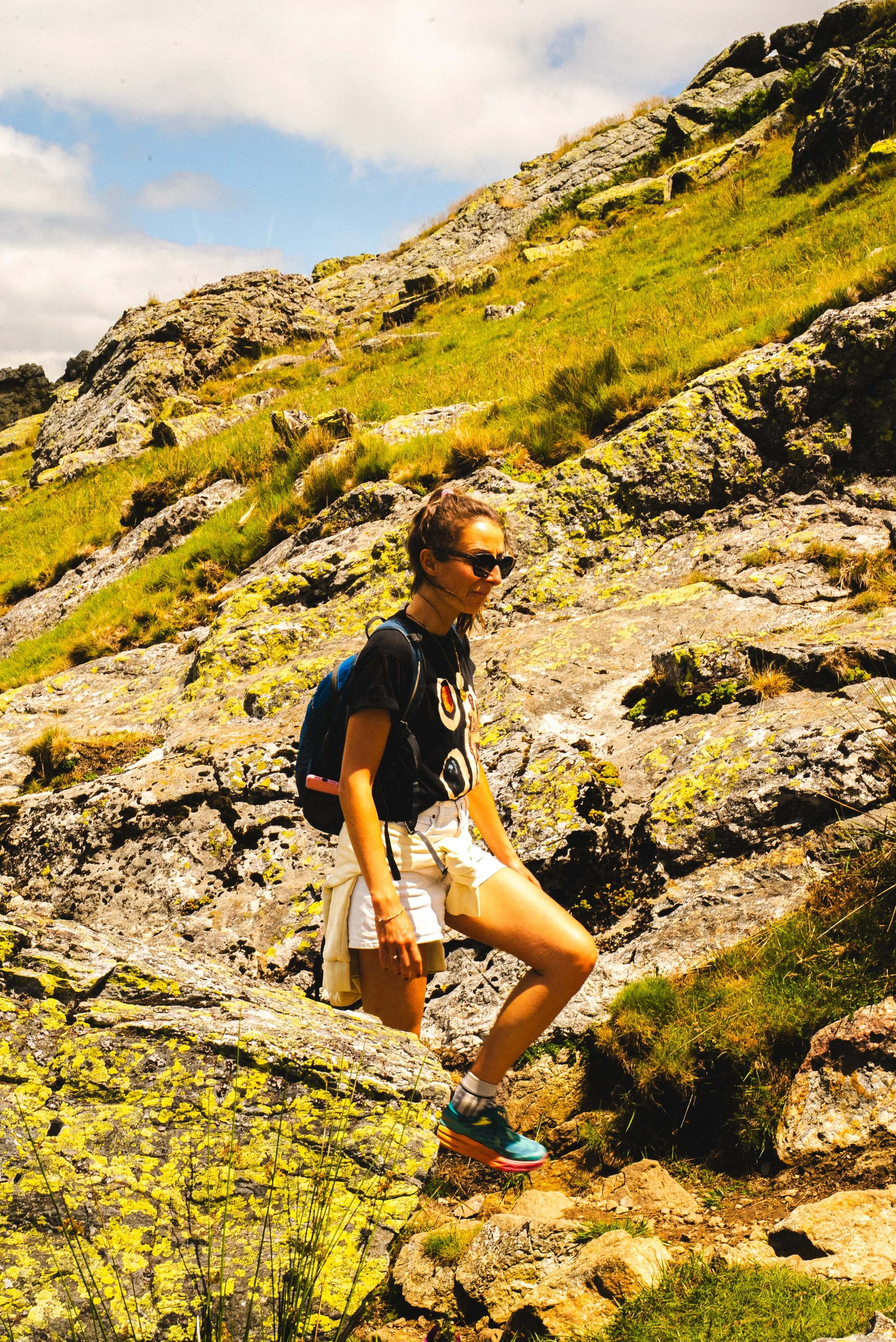 Une femme en randonnée dans un paysage de montagnes rocheuses et verdoyantes, portant une tenue sportive et un sac à dos.