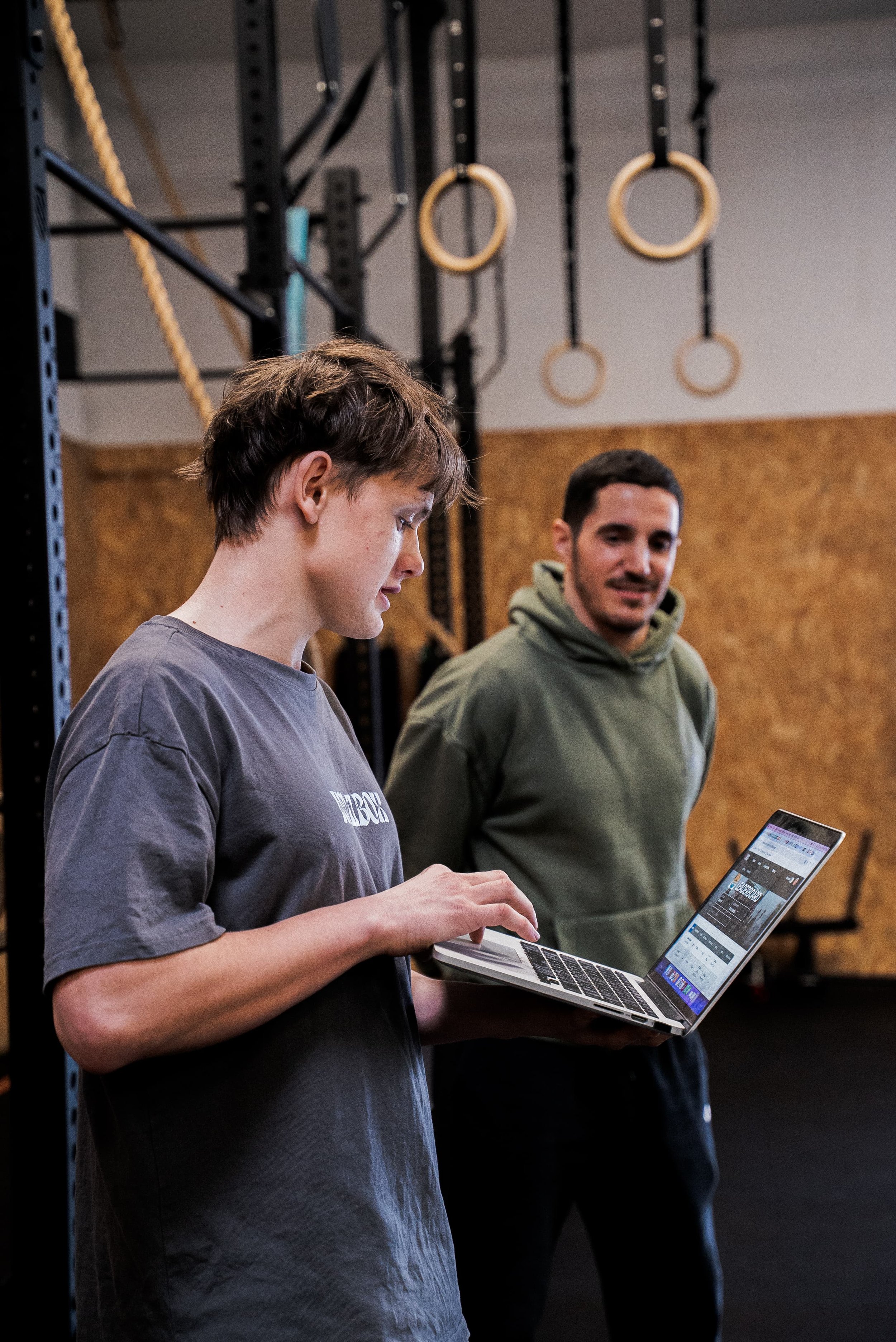 Une femme regarde un ordinateur portable dans une salle de sport ou une salle d'entraînement, avec un homme debout à côté, qui regarde avec un sourire. En arrière-plan, il y a des anneaux de gymnastique suspendus au plafond.