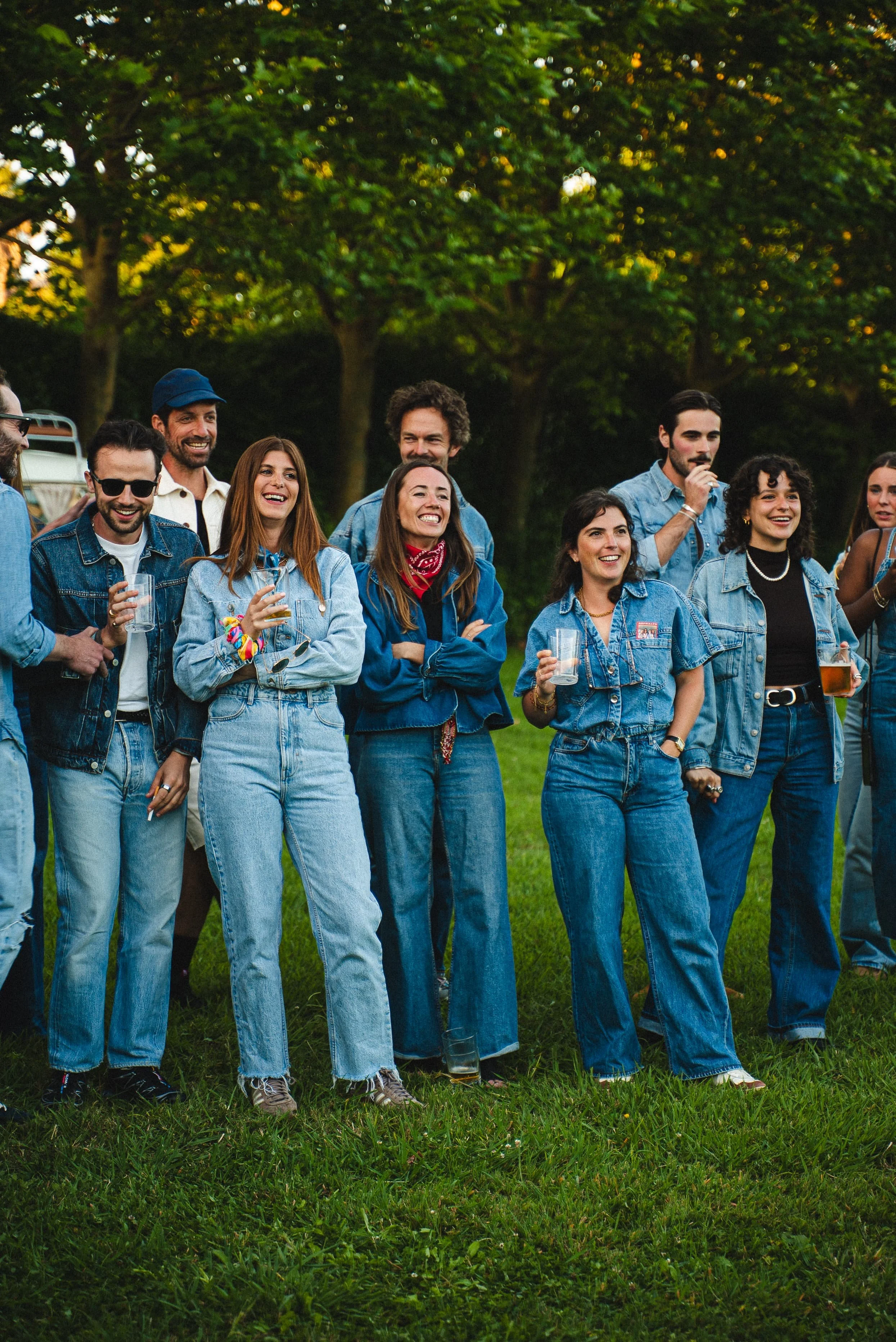Groupe de personnes souriantes et détendues réunies dans un parc, portant des vêtements en denim, lors d'une soirée ou d'une fête en plein air.