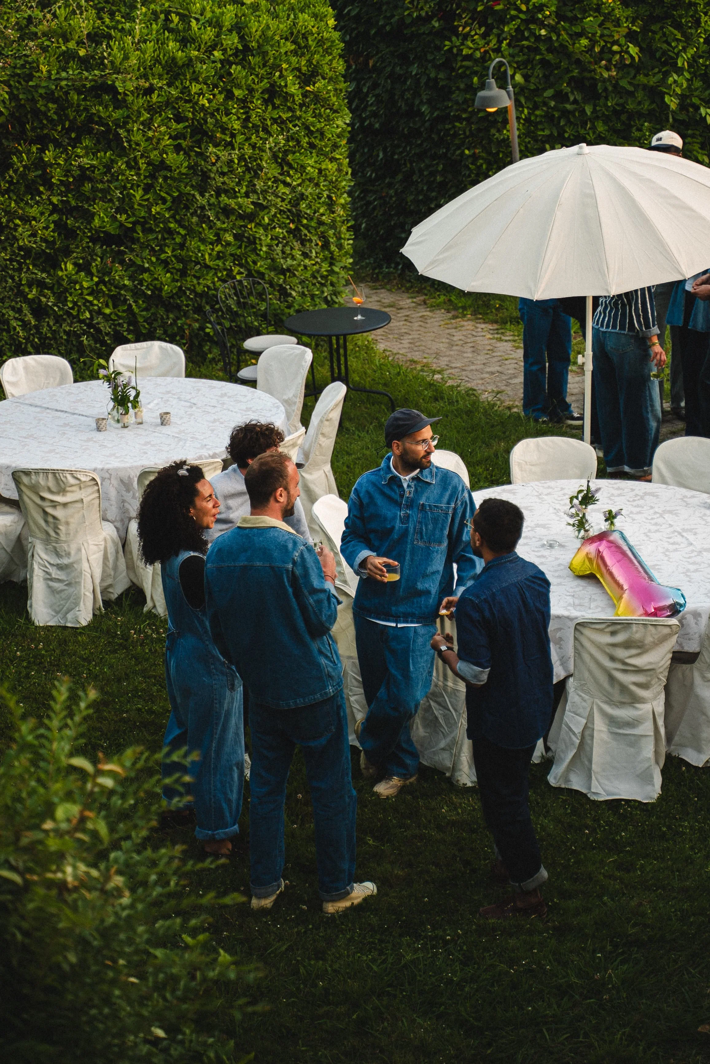 Groupe de personnes lors d'une fête en extérieur, autour d'une table avec des chaises recouvertes de housses blanches, sous un grand parasol blanc. Il y a des boissons sur la table, et des invités parlent et boivent. La scène est dans un jardin avec 