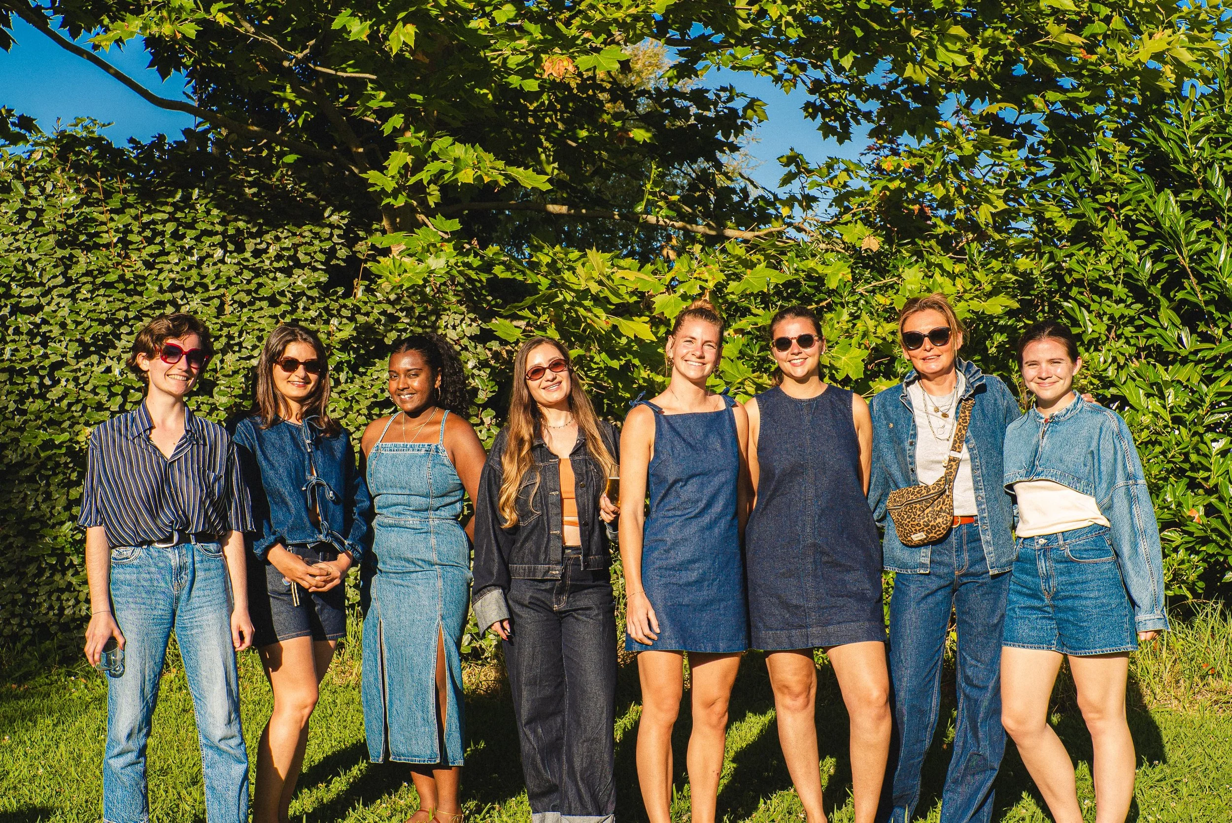 Groupe de huit femmes souriantes posant ensemble dans un espace extérieur ensoleillé, avec des vêtements décontractés en jean et lunettes de soleil, devant un écran de verdure composée de feuillage.