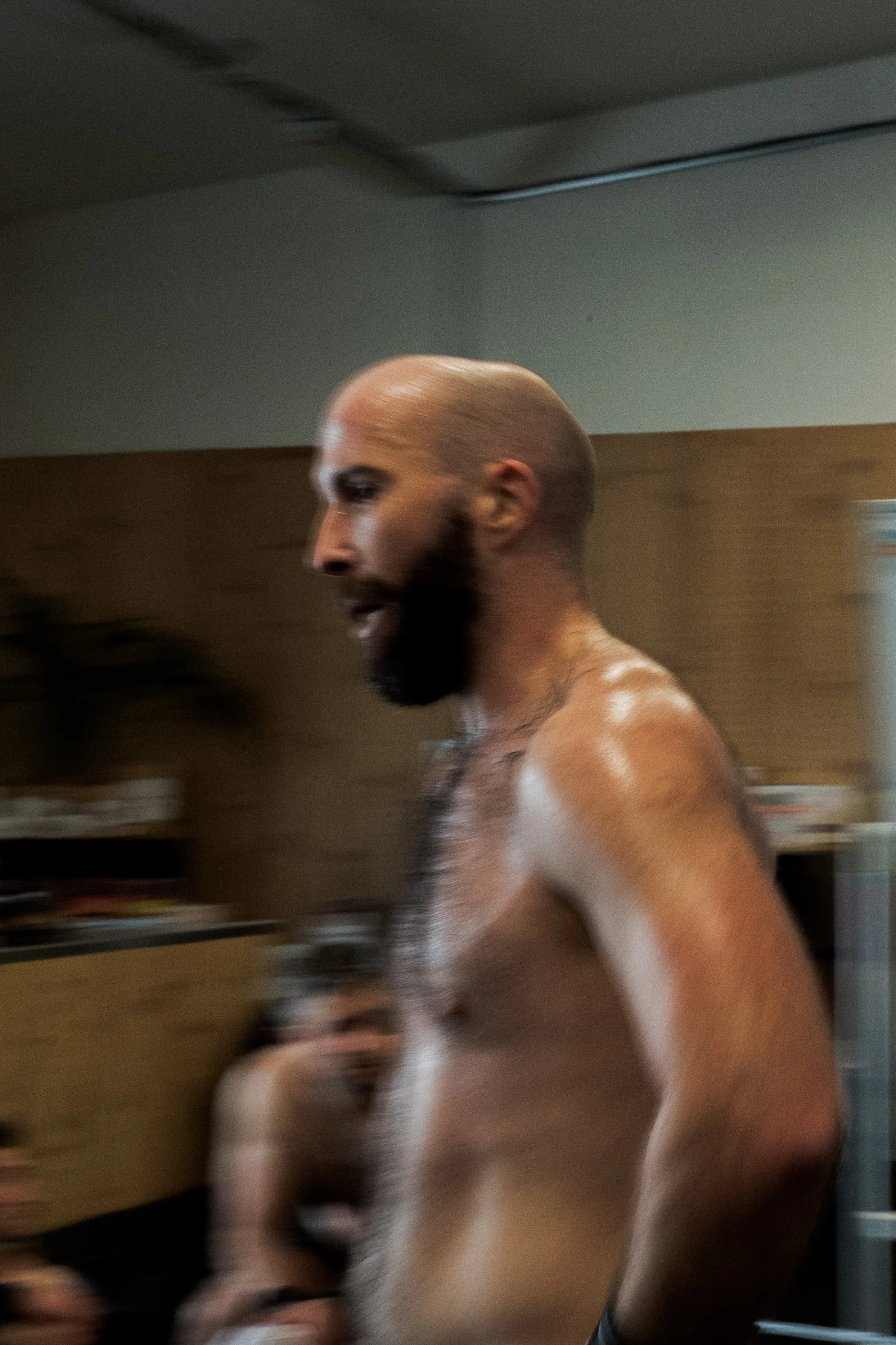 Un homme torse nu, avec un crâne rasé et une barbe foncée, est en mouvement dans un intérieur, flou en raison du mouvement.