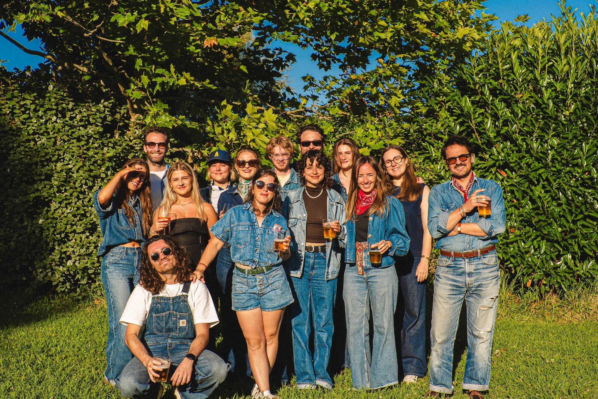 Groupe de personnes posant dehors sous un arbre avec un ciel bleu, portant des vêtements en denim et tenant des verres de bière, souriant et profitant du soleil.