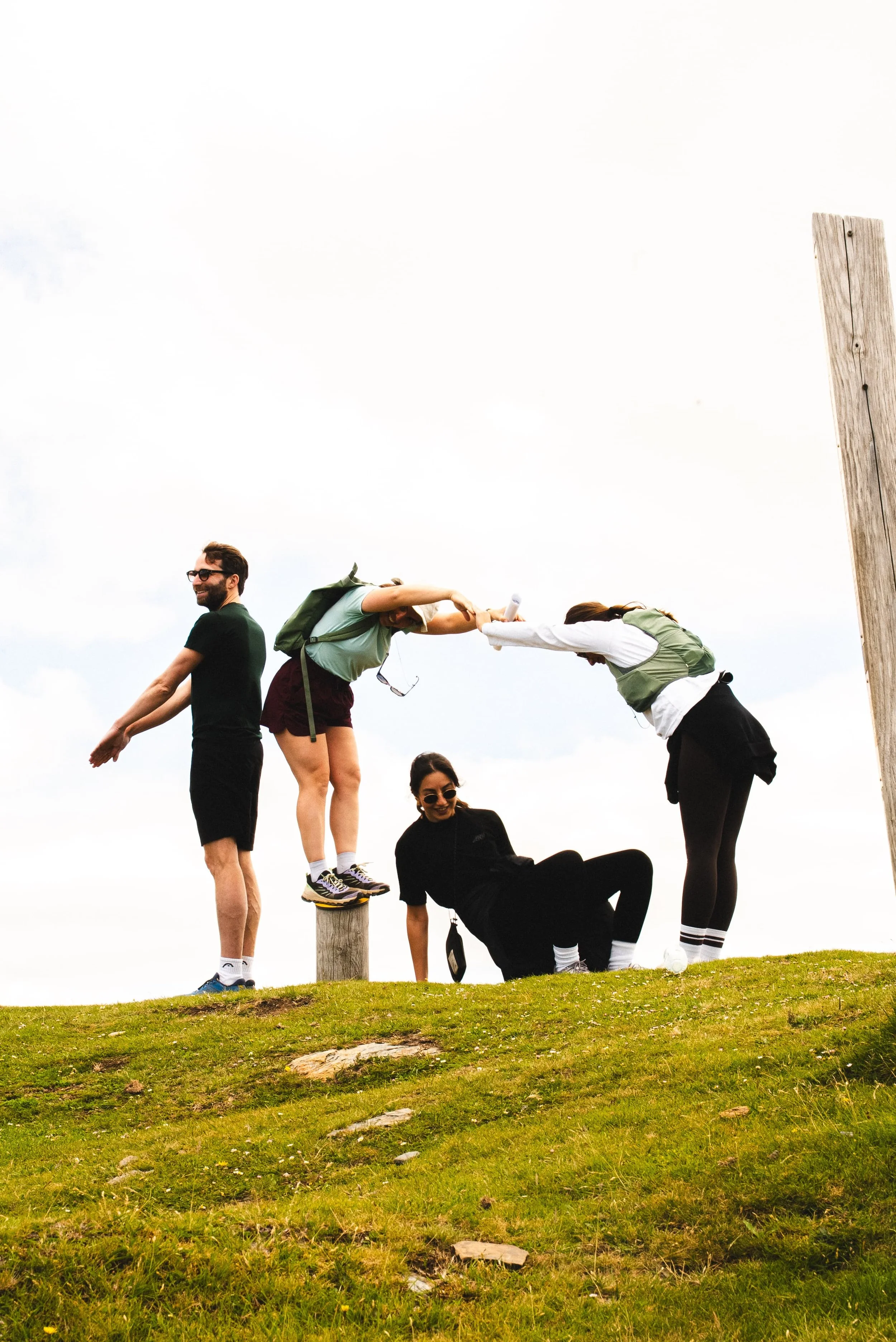 Groupe de cinq personnes posant sur une colline verte avec un panneau en bois, certains debout sur un poteau, d'autres assis par terre, toutes souriantes, sous un ciel clair.