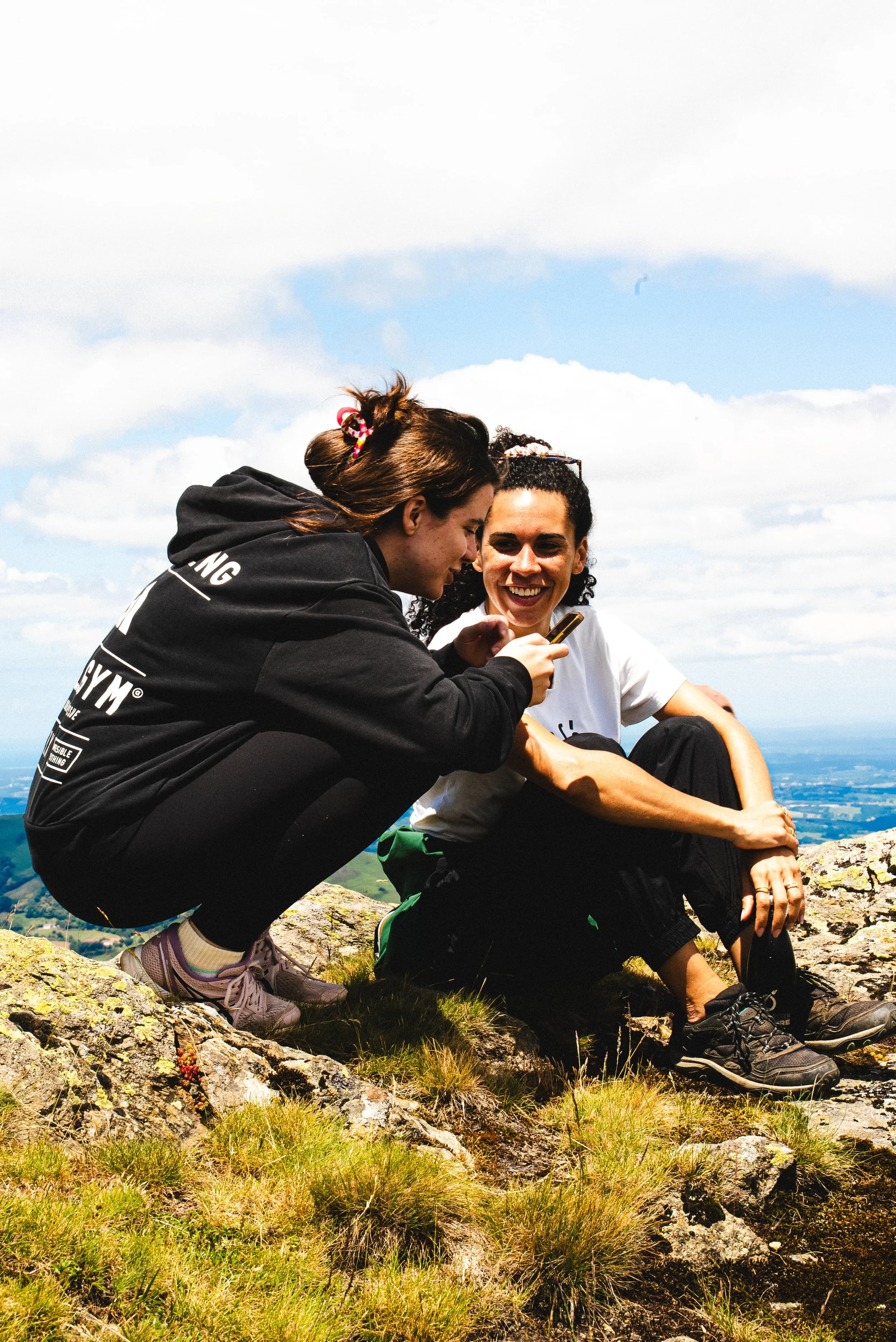 Deux femmes blanches assises sur un rocher de montagne souriantes, l'une utilisant un téléphone portable, avec un ciel partiellement nuageux.