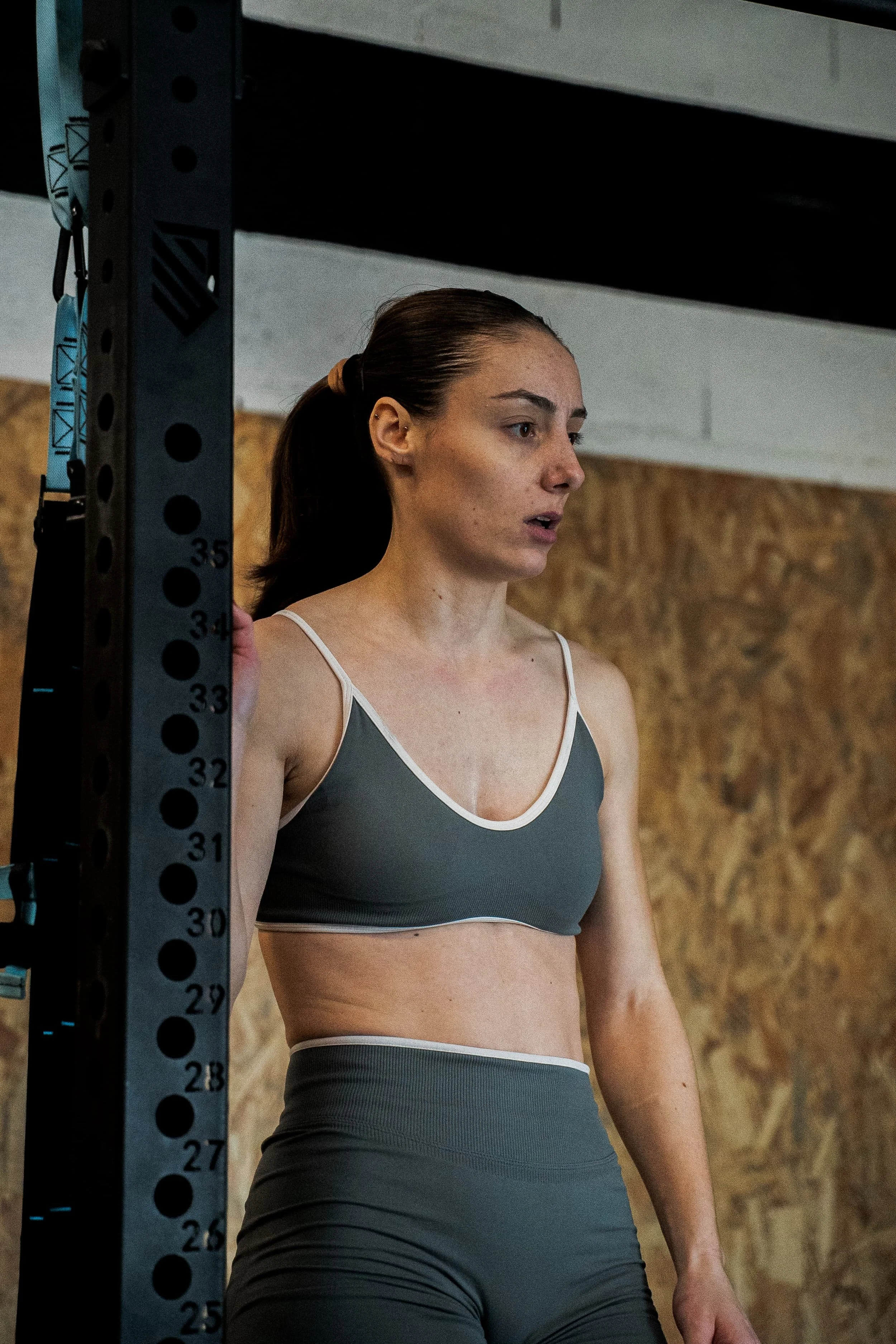 Une femme en tenue de sport à côté d'une structure de fitness en salle, regardant vers la droite, avec un mur en bois en arrière-plan.