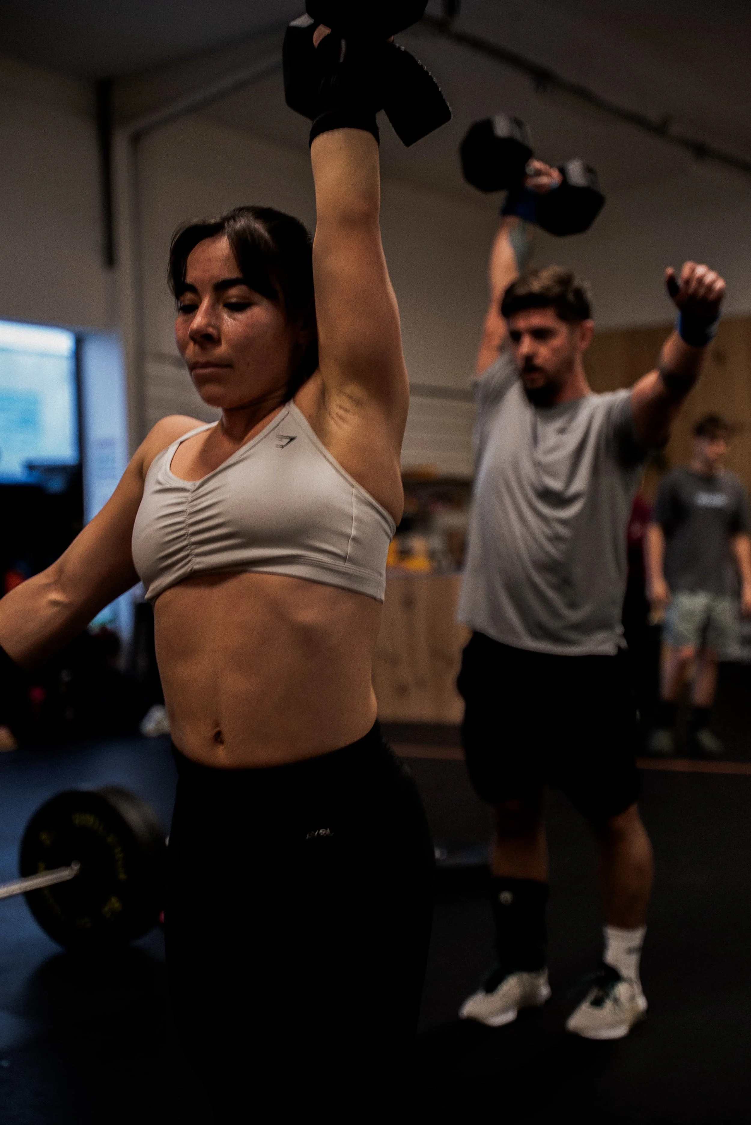 Une femme effectue un exercice de musculation avec un haltère dans une salle de sport, avec un homme derrière qui augmente la charge de haltère.