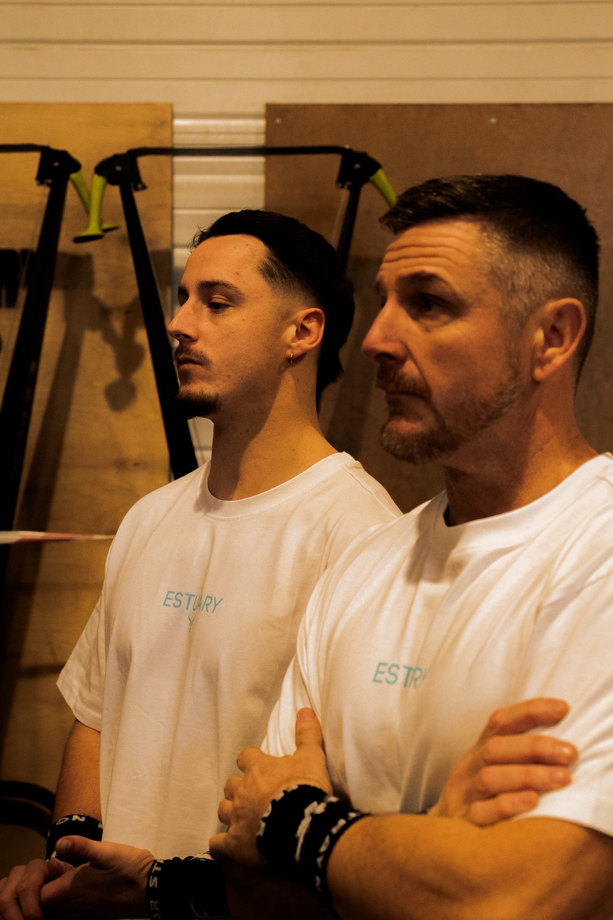 Deux hommes debout côte à côte, portant des t-shirts blancs, dans une pièce avec des murs en bois. Un bras avec un bracelet noir et blanc, probablement d'une femme, est visible en premier plan.