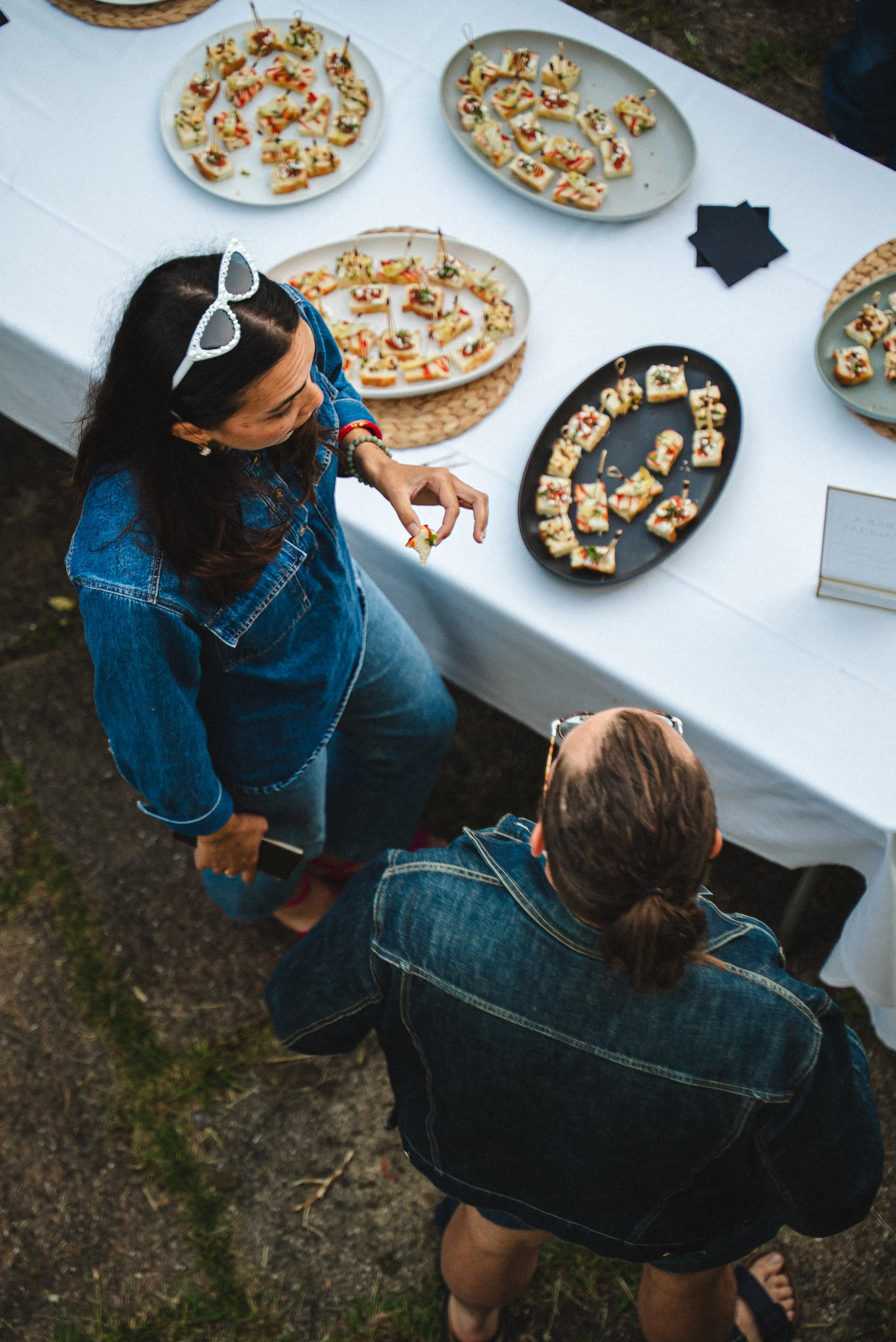 Vue du haut d'une table avec des assiettes de mini sandwiches ou canapés, une personne en denim, deux autres personnes en denim, vue de dessus, en extérieur.