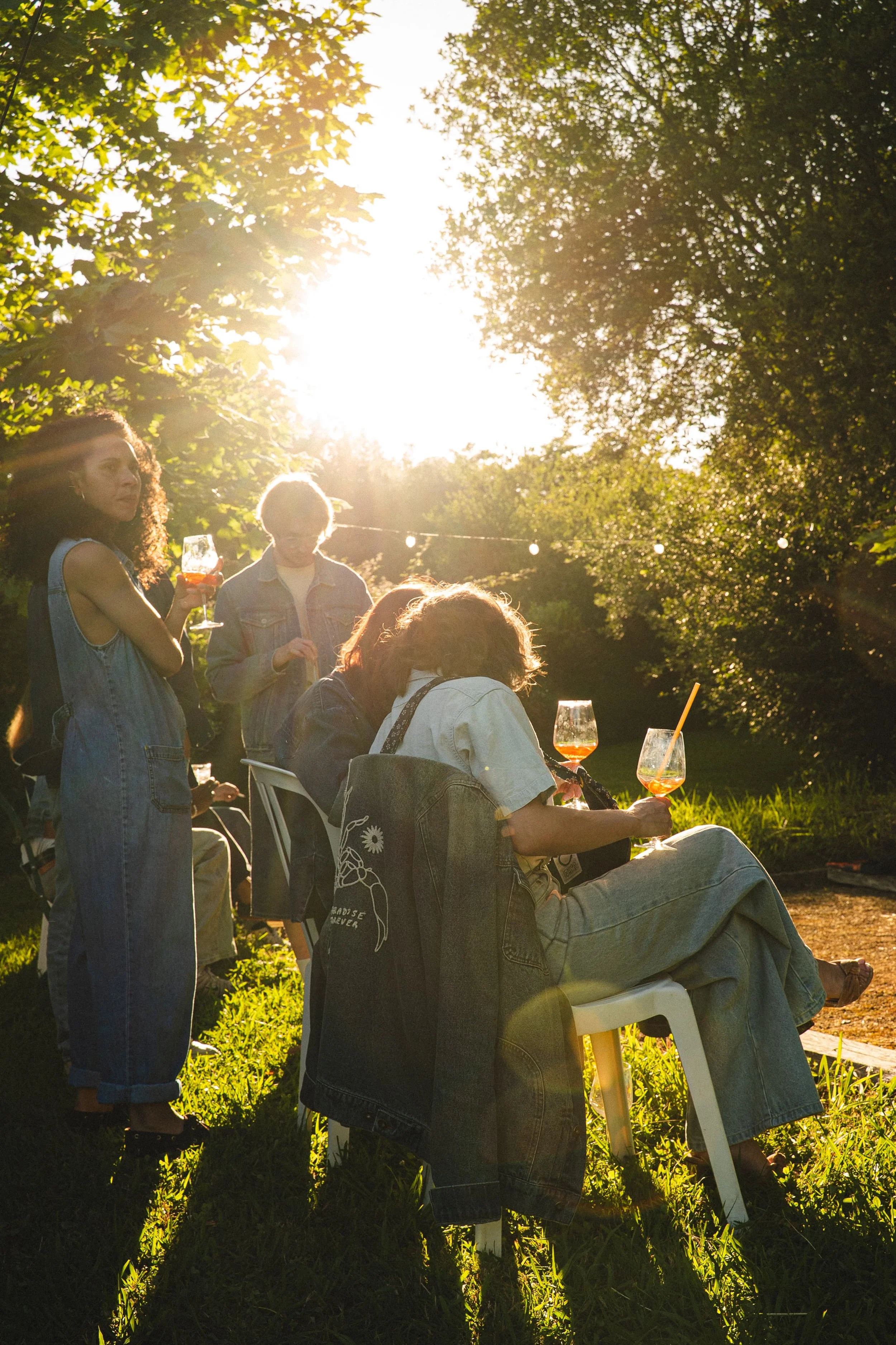Groupe de jeunes assis et debout lors d'une réunion en plein air en soirée, entouré d'arbres, tenant des boissons colorées.