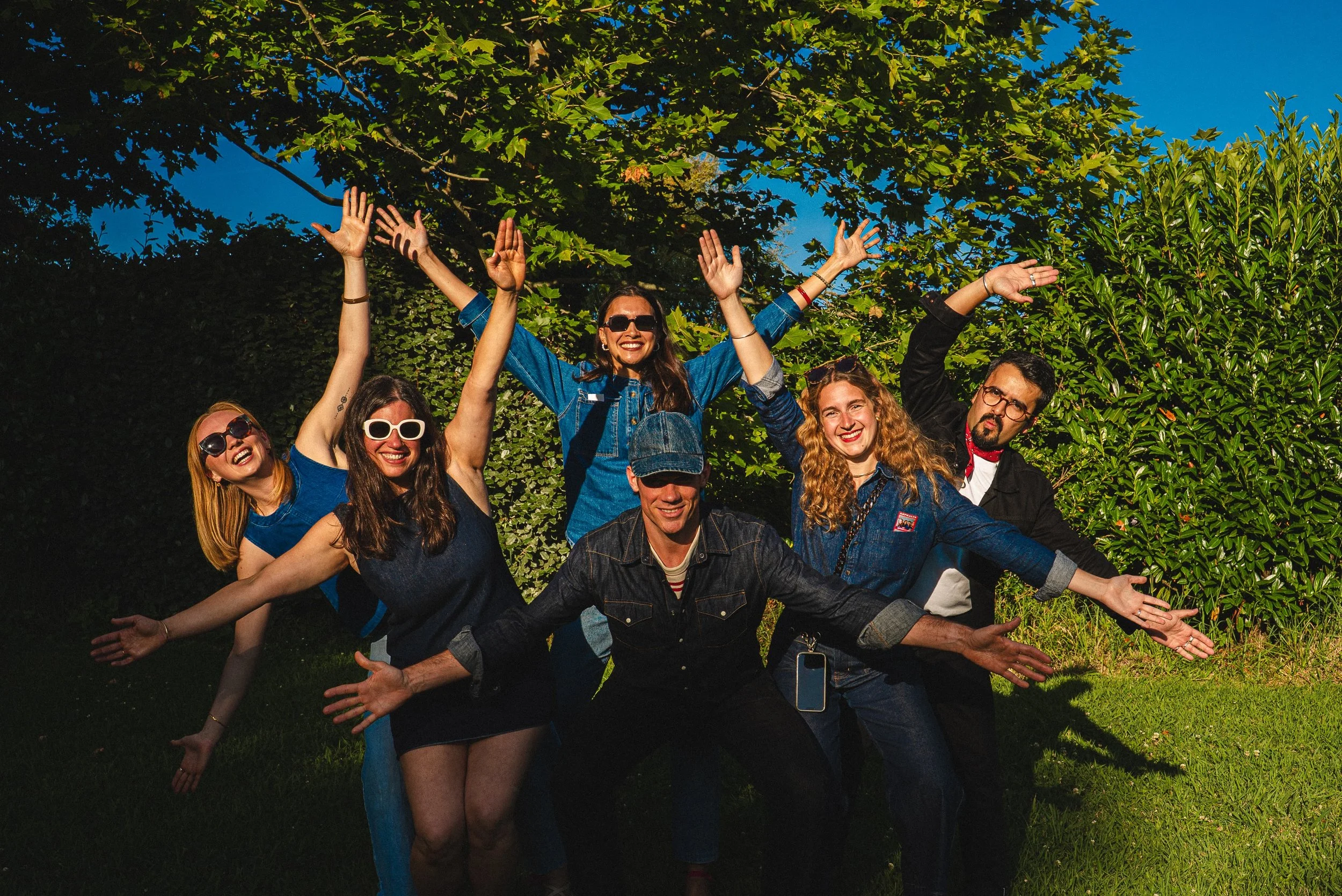 Groupe de personnes souriantes posant en forme d'étoile avec les bras levés dans un parc ensoleillé, arrière-plan d'arbres verts et ciel bleu.