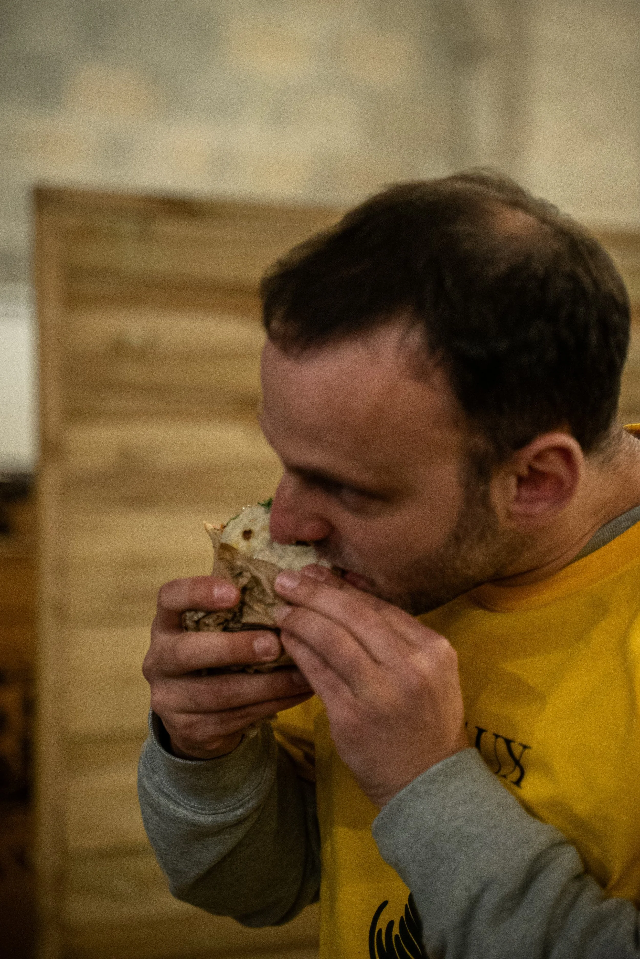 Un homme mange un sandwich avec ses mains, portant un sweat à manches longues gris et un t-shirt jaune.