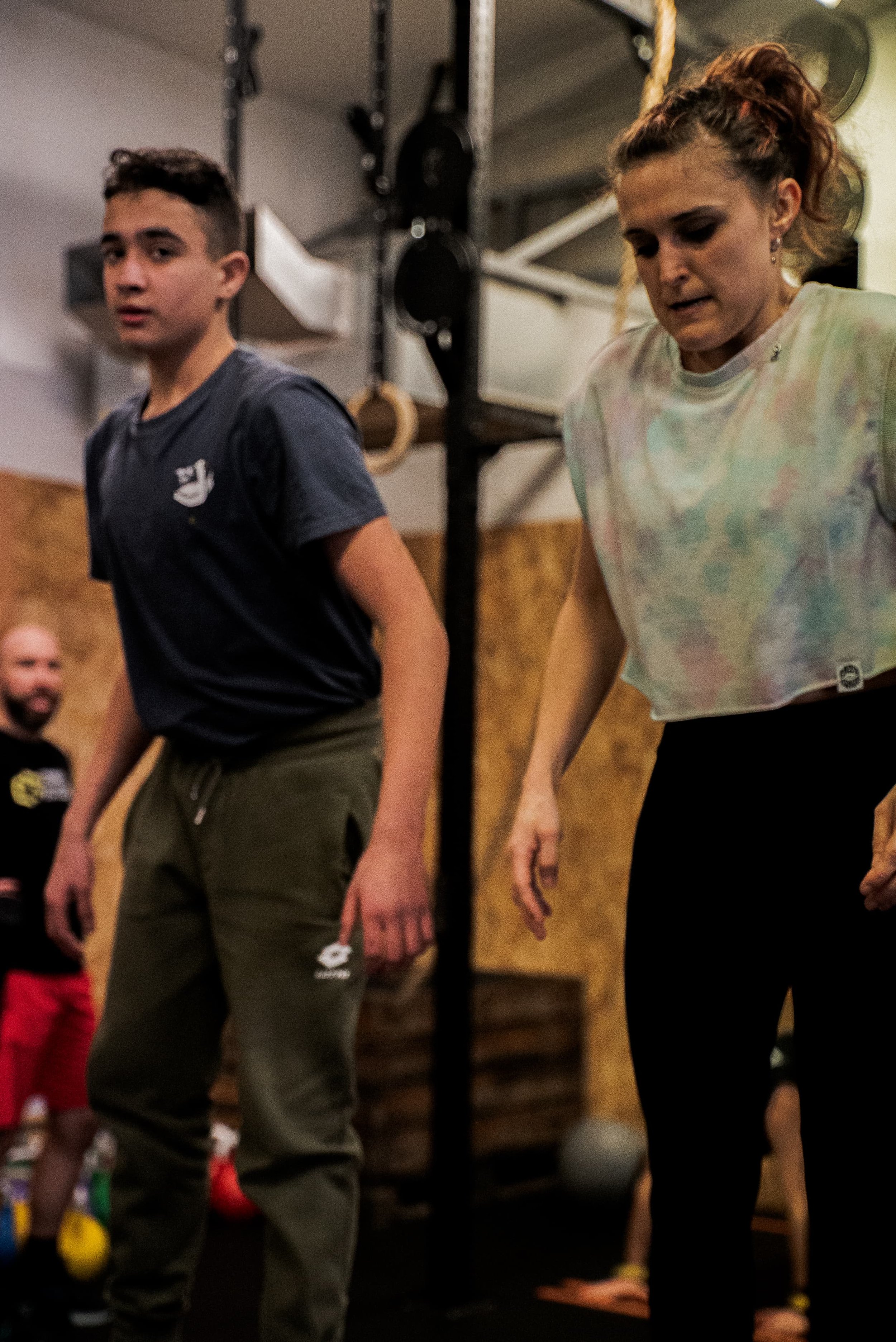 Deux personnes en train de faire un exercice physique dans une salle de sport, avec un fond de matériel d'entraînement. Une femme regarde vers le bas, l'autre personne est légèrement floue.