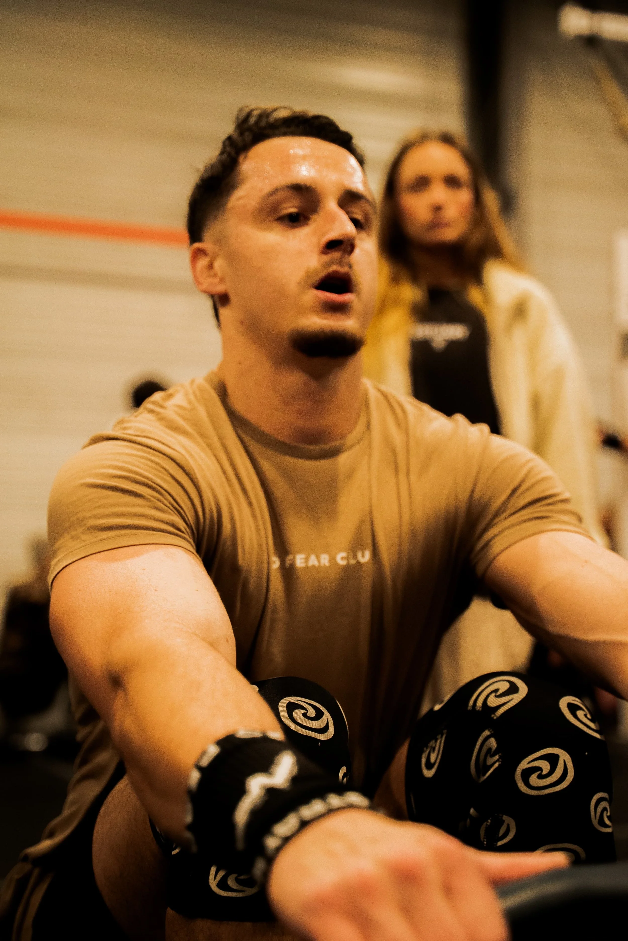 Un homme en tenue de sport portant des bandes de boxe, concentré lors d'une séance d'entraînement en salle de sport. En arrière-plan, une femme observe.