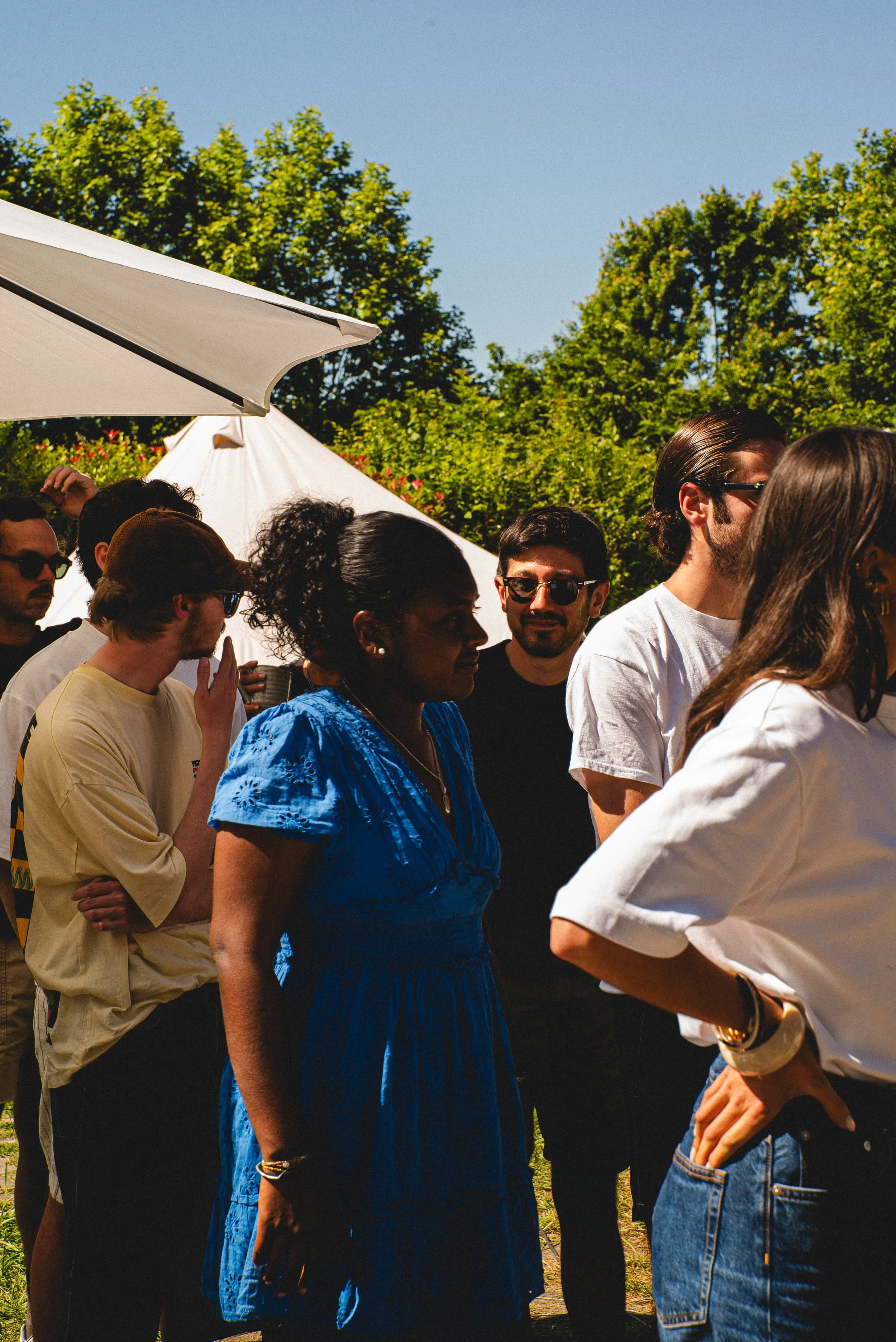 Groupe de personnes à l'extérieur, sous un parasol blanc, avec des arbres en arrière-plan, en pleine journée ensoleillée.