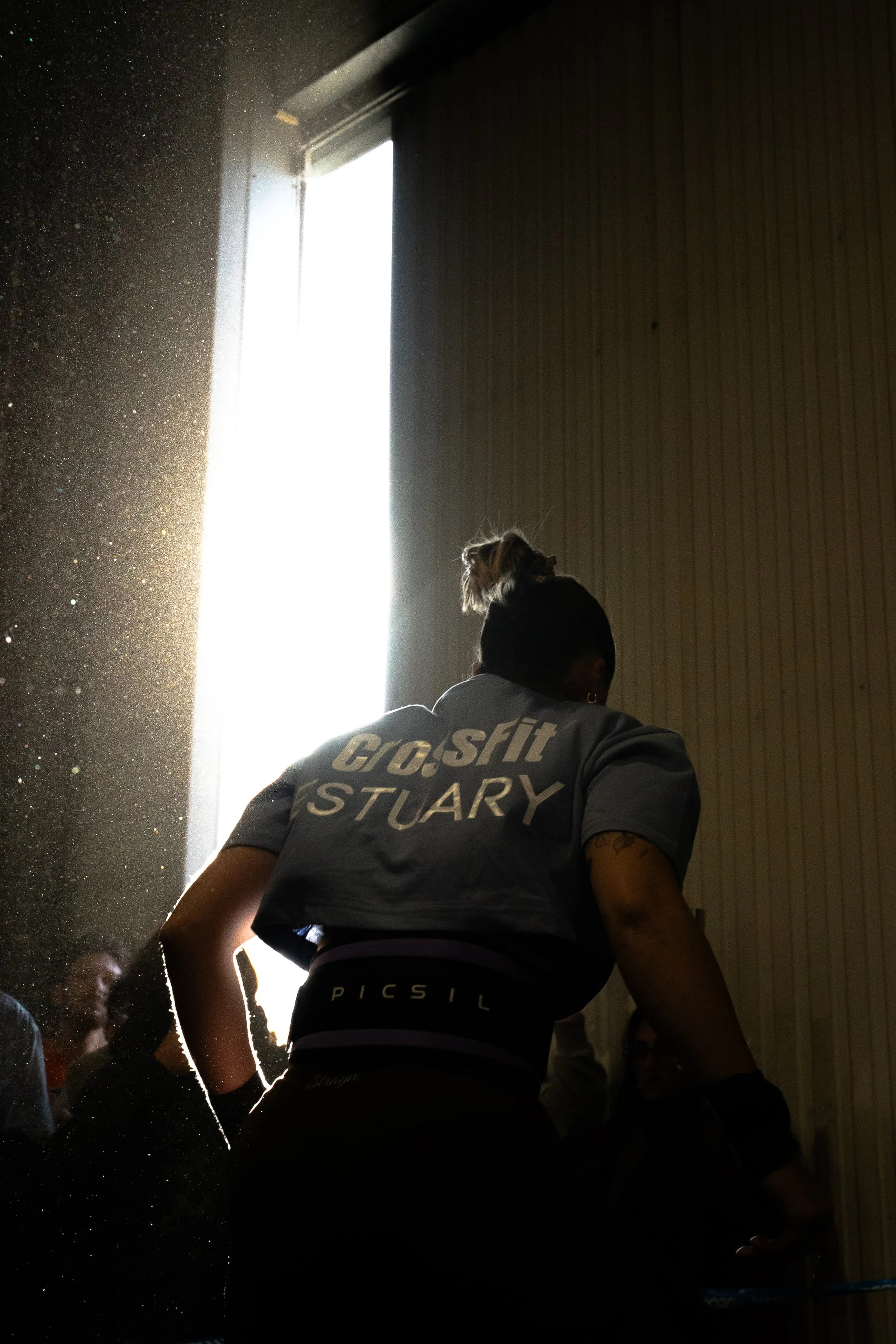 Une femme portant un t-shirt gris avec des inscriptions visibles tourne le dos, et un lion en peluche est placé sur sa tête. La scène est éclairée par une source lumineuse forte derrière elle, créant un contraste intense et une ambiance sombre à l'intérieur.