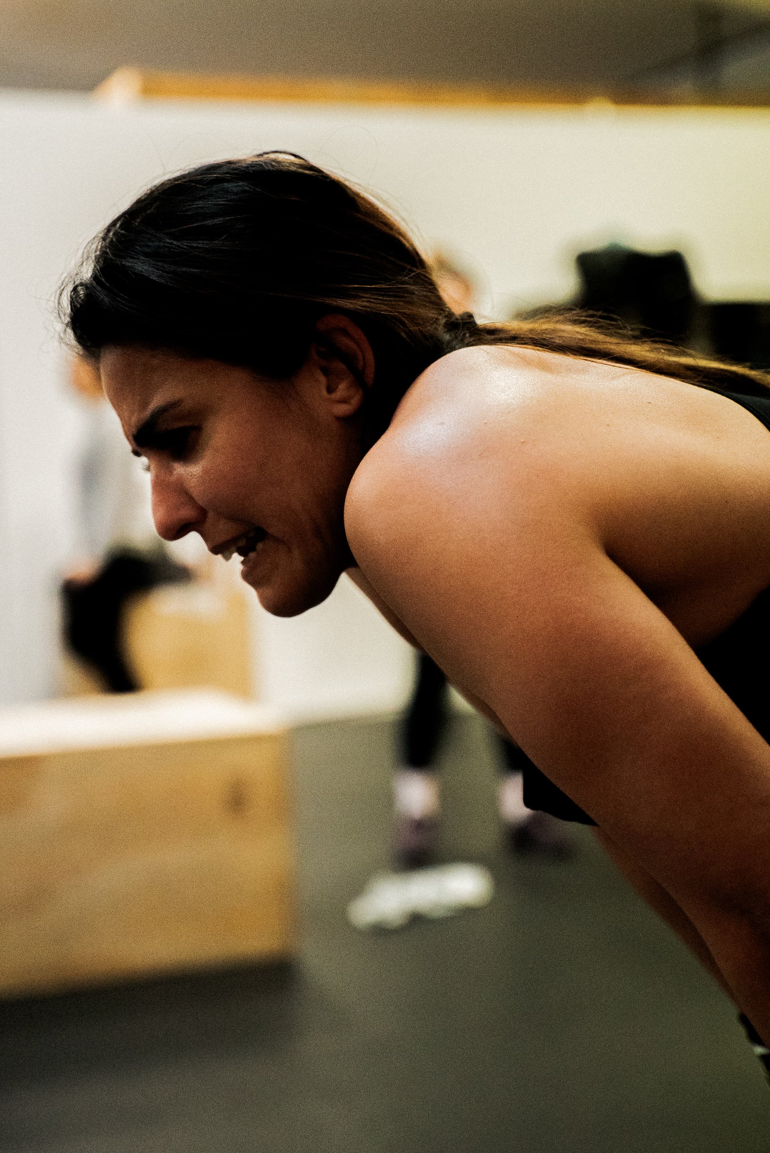 Une femme souriante avec des cheveux bruns foncés, en train de faire du sport ou de s'entraîner, dans un environnement intérieur, probablement une salle de sport.