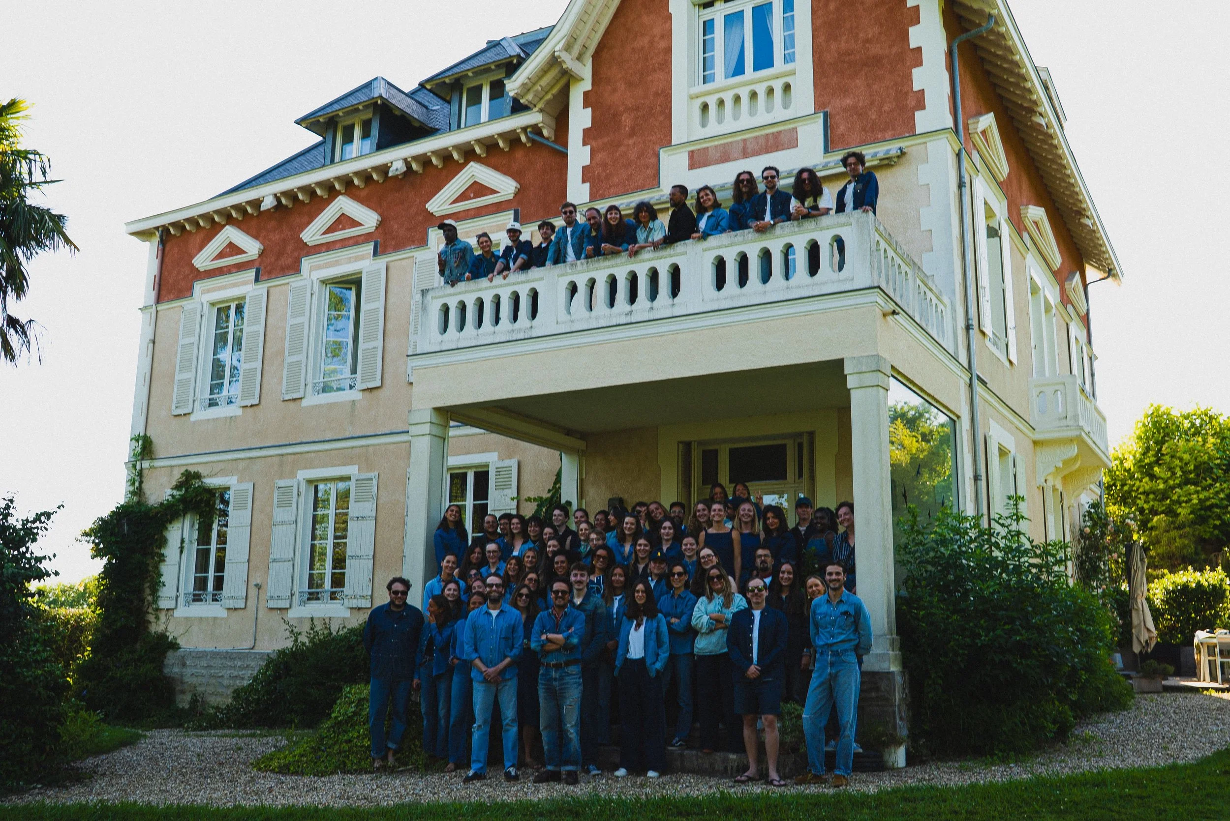 Un grand groupe de personnes posant devant une grande maison colorée. La maison a plusieurs fenêtres avec des volets blancs et un balcon dans la partie supérieure. Le groupe est composé d'hommes et de femmes souriants, certains portant des lunettes, 